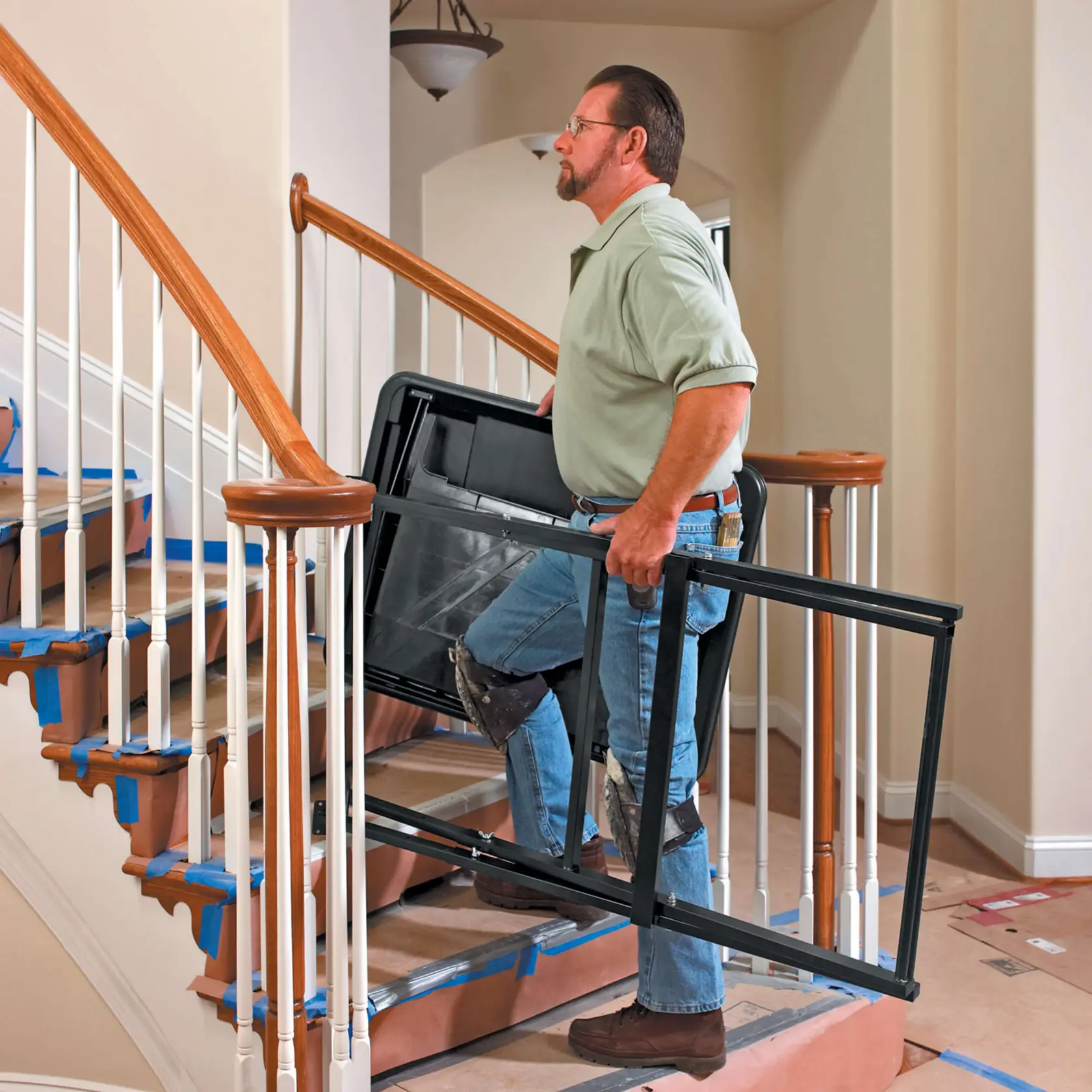 A person wearing knee pads carries a DEWALT D24000 tile saw up a staircase. The stairs are covered with protective paper and blue tape. The setting appears to be indoors during a renovation.