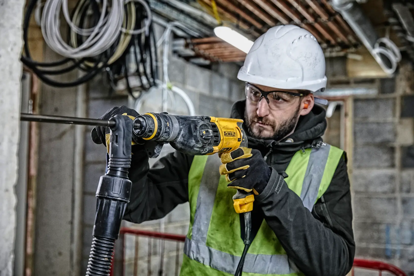 Eine Person mit Sicherheitshelm, Handschuhen und Warnweste verwendet eine DEWALT D25134K Bohrhammer auf einer Baustelle. Im Hintergrund sind sichtbare Kabel und Rohrleitungen in einem unfertigen Gebäude zu sehen.
