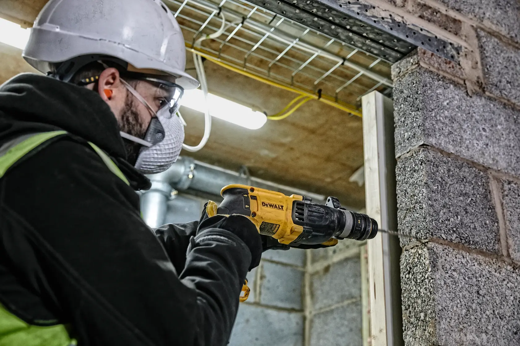 Ein Arbeiter mit Sicherheitshelm und Handschuhen verwendet einen DEWALT D25134K Bohrhammer, um eine Betonwand auf einer Baustelle zu bohren.