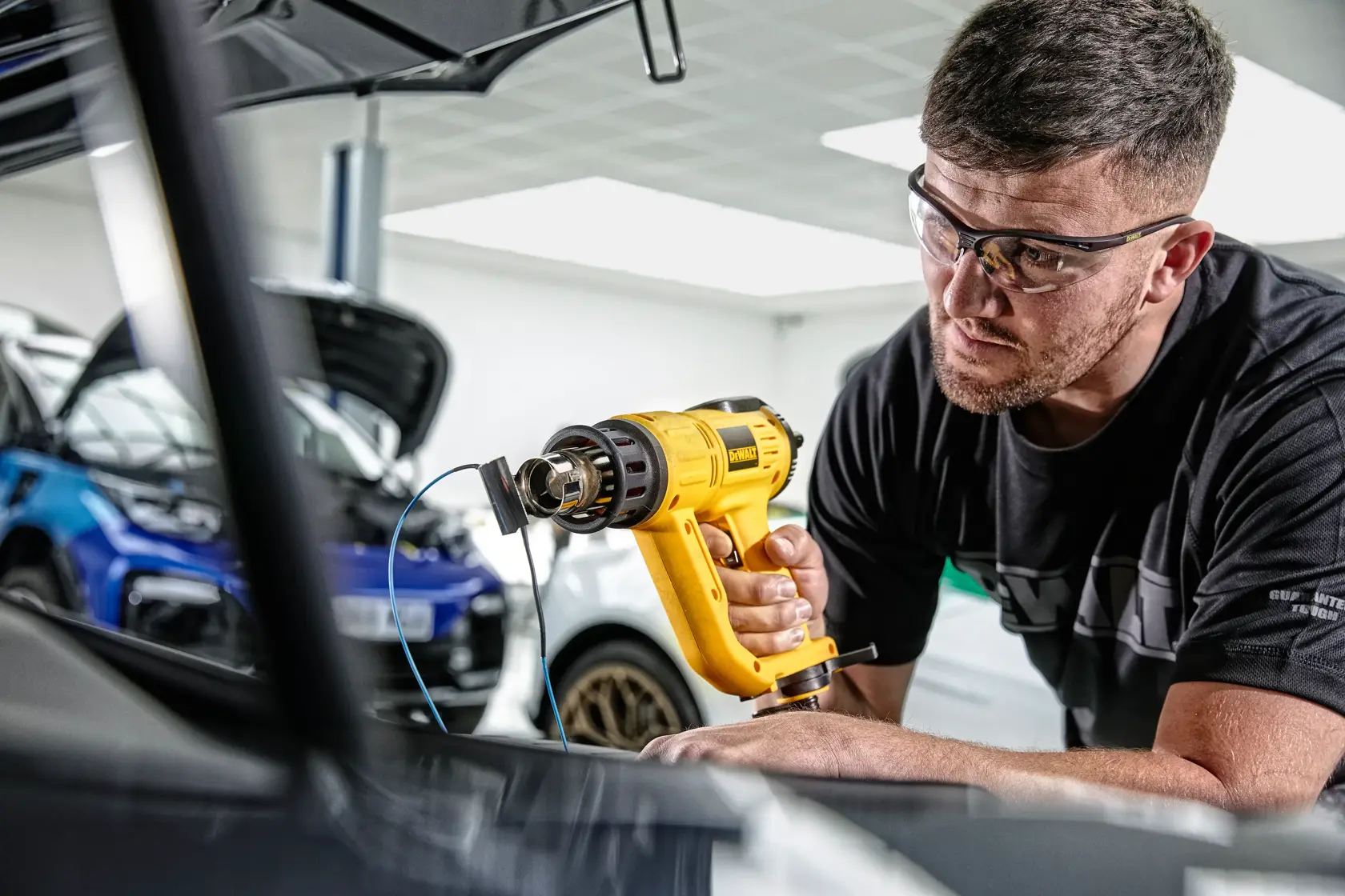 Una persona utiliza una pistola de calor amarilla DEWALT D26414 en un garaje o taller, con coches visibles al fondo. El rostro de la persona está difuminado para mantener la privacidad.