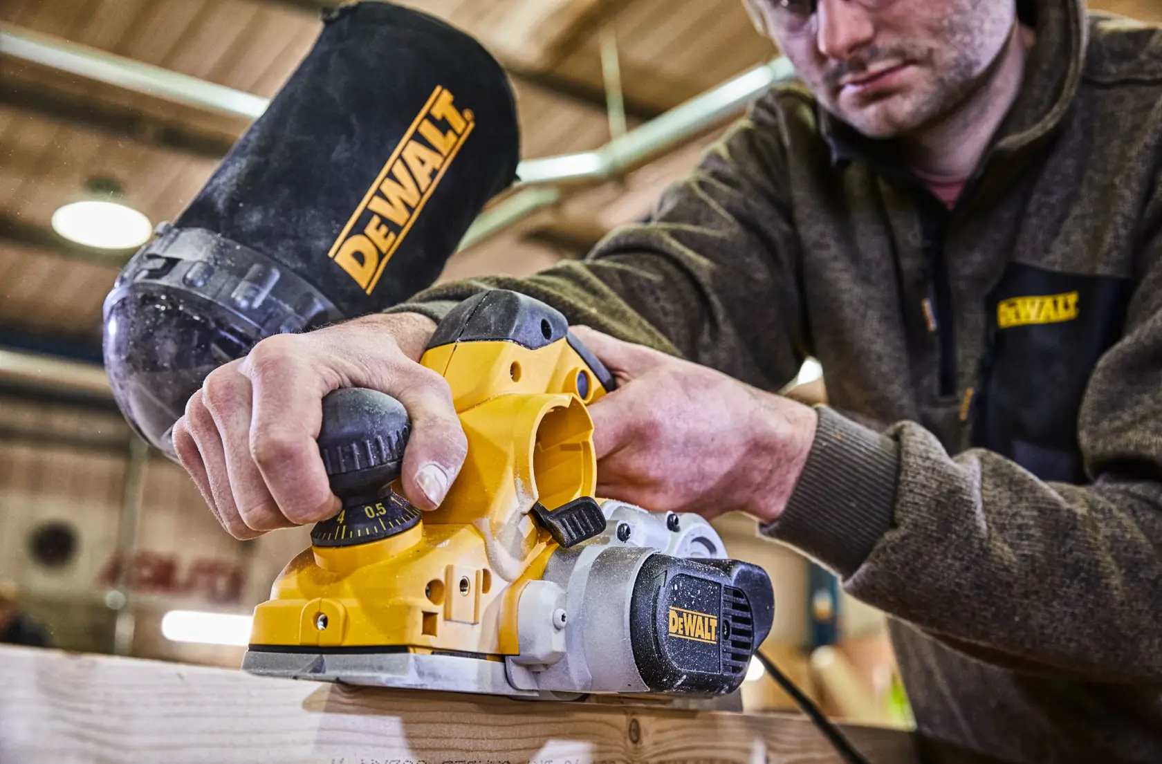Una persona utiliza una cepilladora eléctrica DEWALT D26500 sobre una superficie de madera en un taller. El rostro de la persona está difuminado y el logotipo de DEWALT es visible en la bolsa de polvo y en su ropa.
