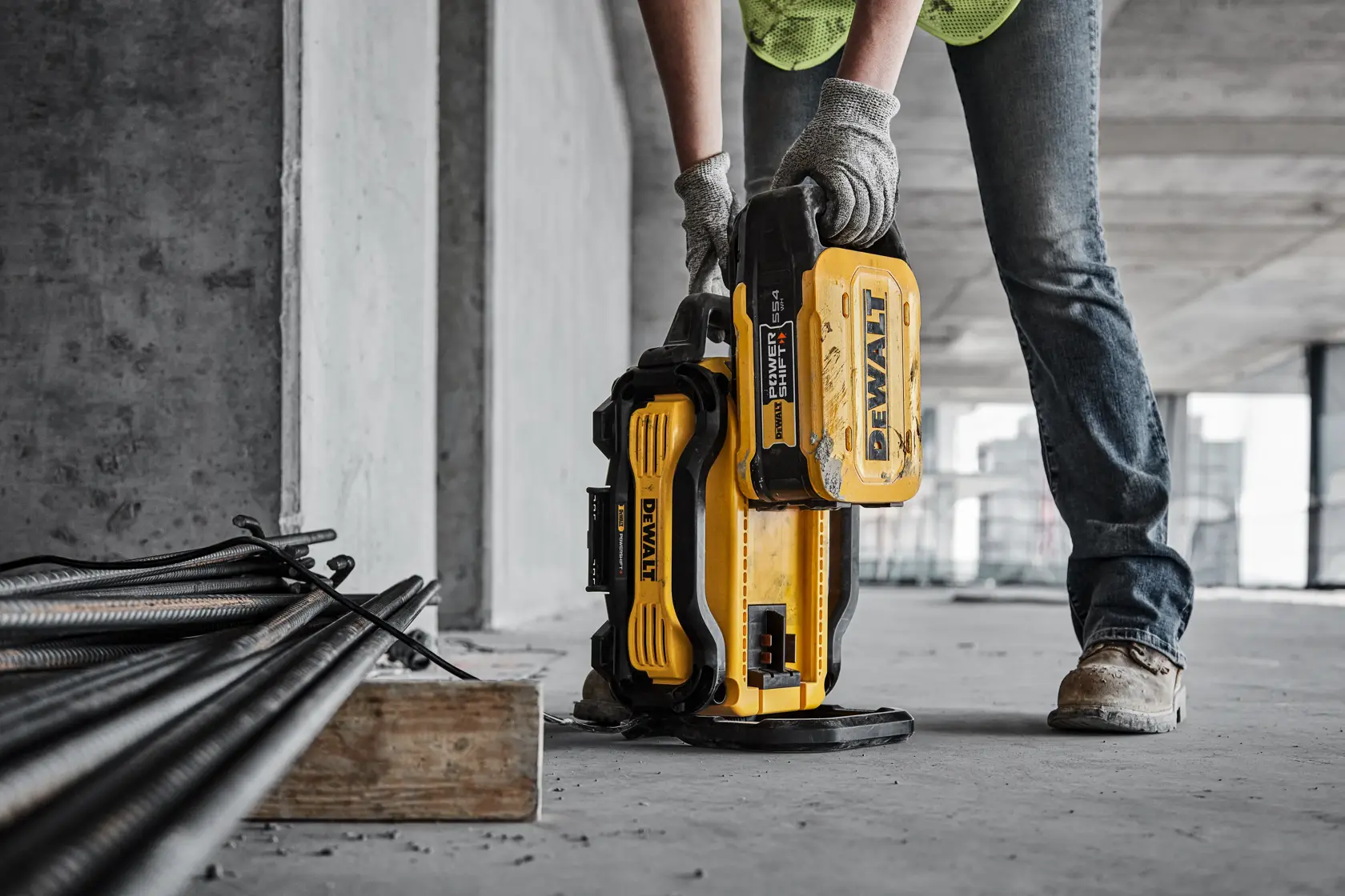 Persona utiliza una batería amarilla DEWALT en una obra, con herramientas y barras metálicas cerca.