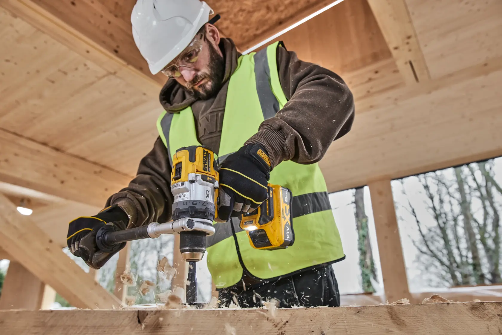 Persona perfora madera con un taladro inalámbrico DEWALT, usando guantes y chaleco de seguridad.