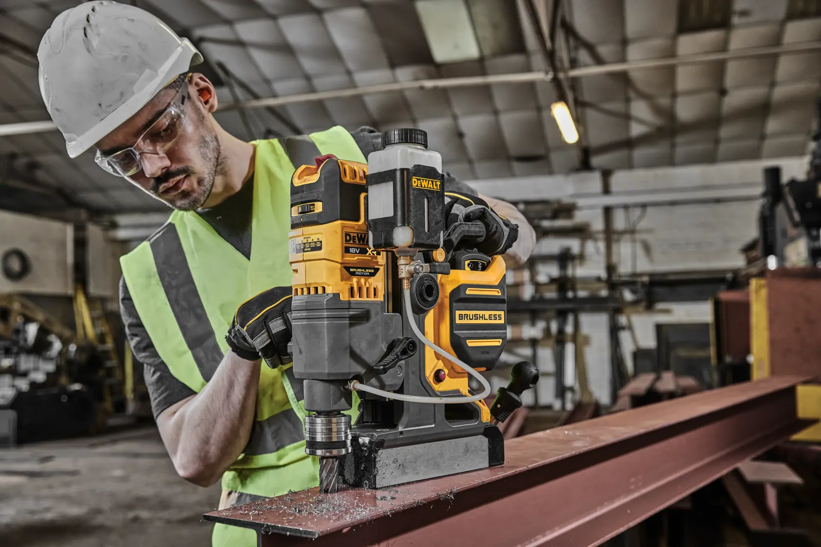 DEWALT brushless magnetic drill in use on a steel beam in an industrial workshop by a worker wearing safety gear.