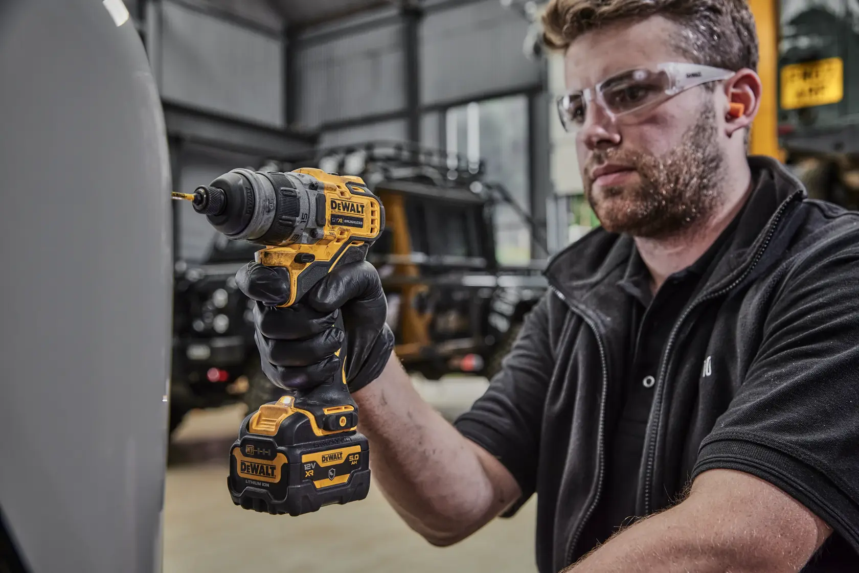 Eine Person mit schwarzen Handschuhen benutzt einen gelben und schwarzen DEWALT-Akkubohrer mit angeschlossenem Akku in einem Innenbereich. Das Gesicht der Person ist aus Datenschutzgründen unscharf gemacht.