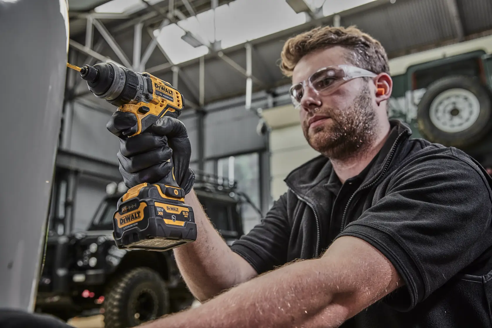 Eine Person mit verschwommenem Gesicht benutzt einen gelb-schwarzen DEWALT DCD703NT Akku-Bohrmaschine in einer Werkstatt oder Garage. Die Person trägt schwarze Handschuhe.