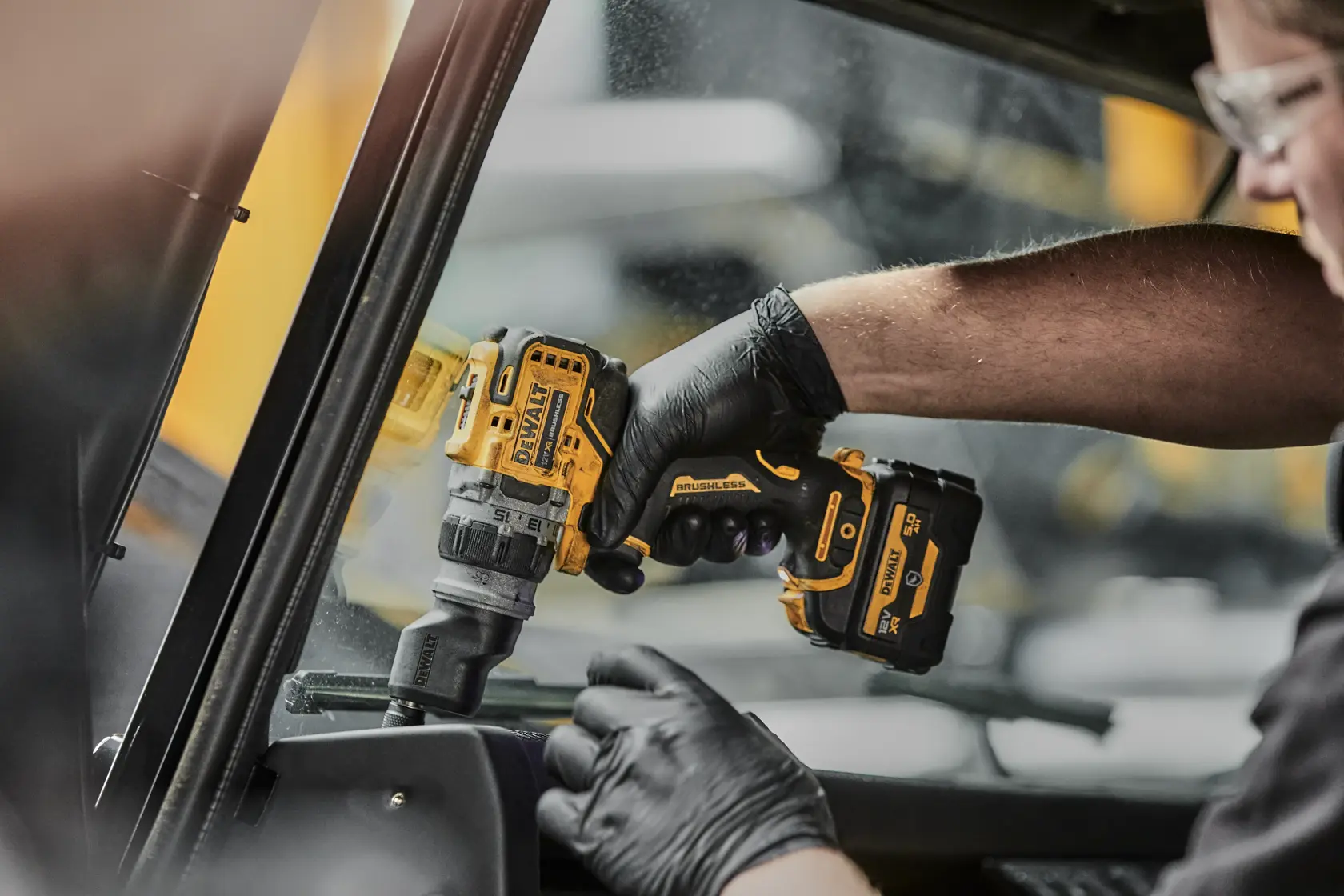 Eine Person mit schwarzen Handschuhen verwendet einen gelb-schwarzen DEWALT-Akkubohrer mit bürstenlosem Motor, um im Inneren eines Fahrzeugs in der Nähe eines Fensters zu arbeiten.