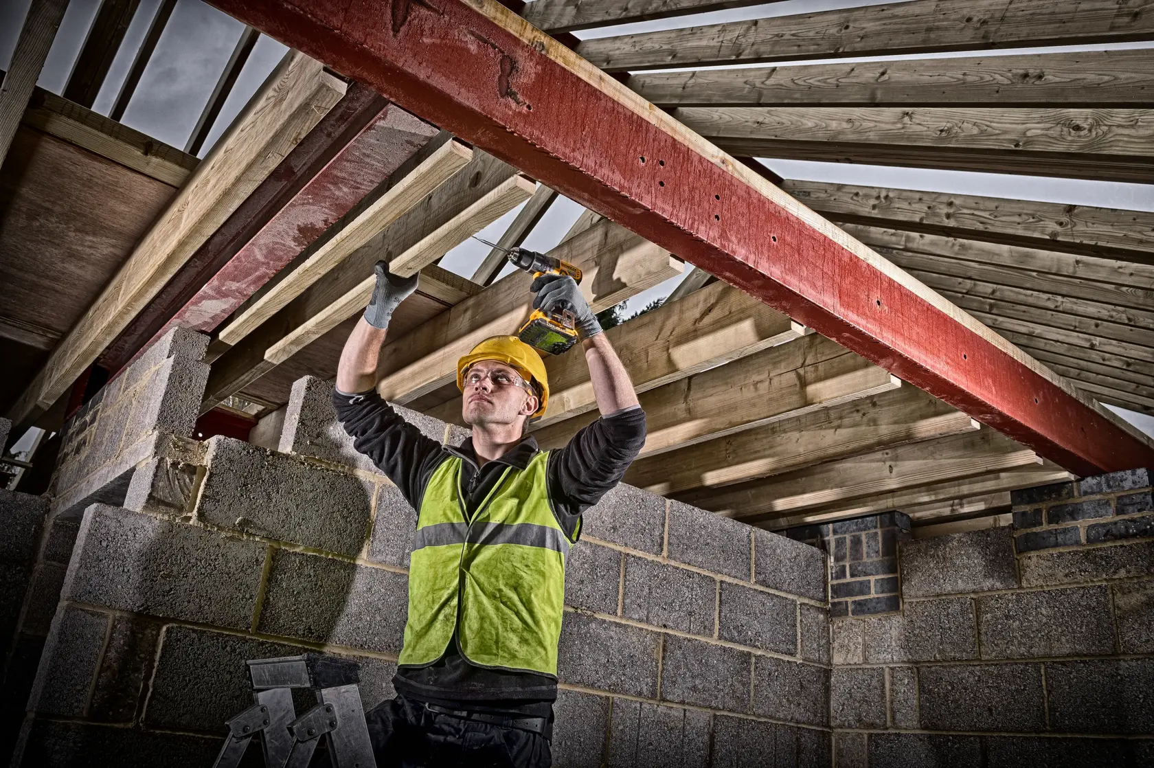 Photo of worker using DEWALT drill DCD791P2 on roof beams at construction site, wearing safety vest and helmet.