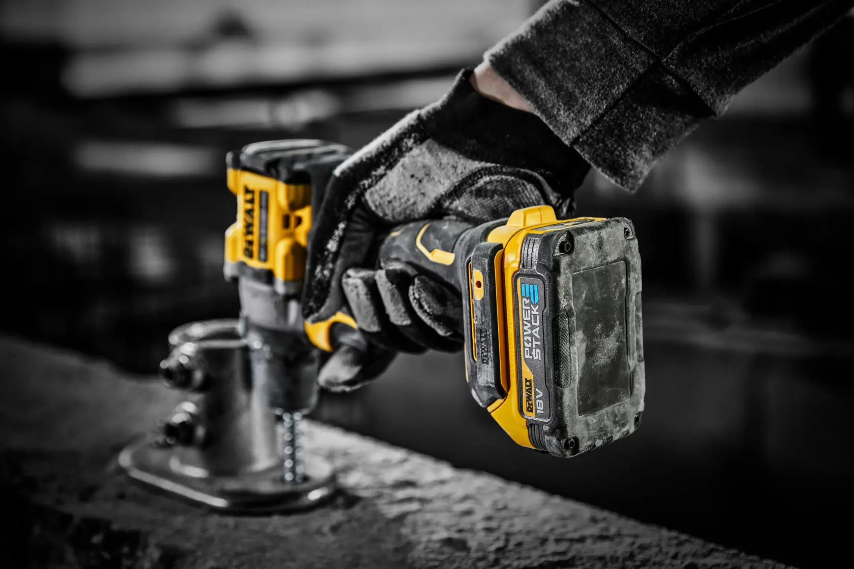 Una mano enguantada sostiene un taladro inalámbrico DEWALT con batería POWERSTACK de 18V, perforando una superficie metálica en un entorno industrial.
