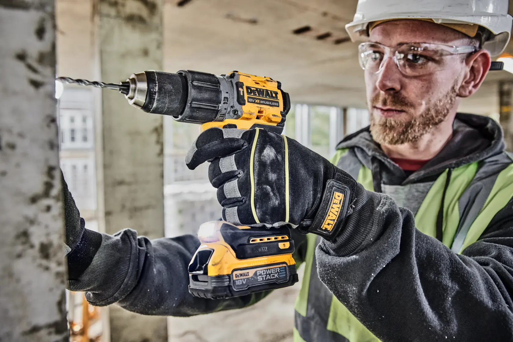Una persona con guantes de seguridad y chaleco reflectante está usando un taladro inalámbrico DEWALT de 18V para perforar una superficie metálica en un sitio de construcción. El taladro es de la marca DEWALT y lleva una batería POWERSTACK.