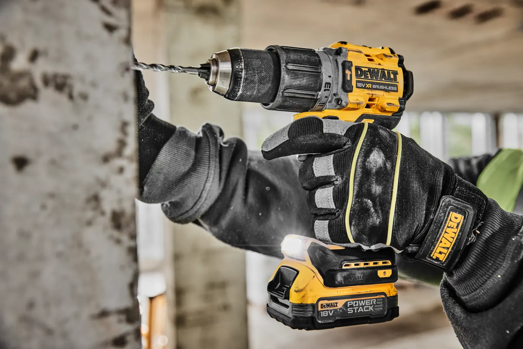 Una persona con guantes de trabajo negros utiliza un taladro inalámbrico amarillo y negro DEWALT para perforar una superficie de concreto en interiores.