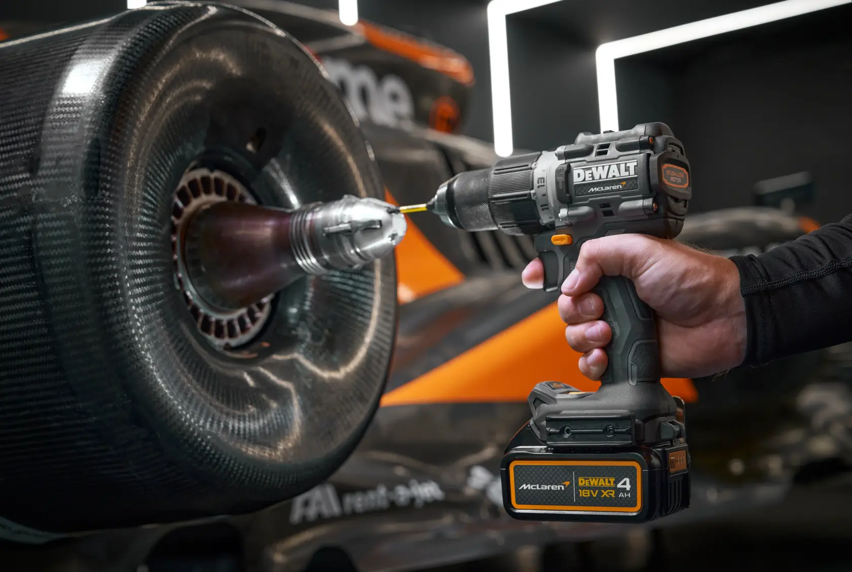 Hand holding DEWALT cordless drill near a race car wheel. Drill is branded McLaren and 18V XR.