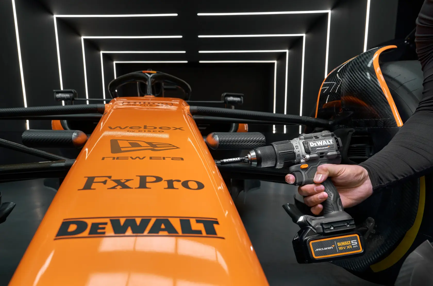 Taladro DEWALT inalámbrico usado en un auto McLaren naranja, ambos con logos de patrocinadores en un garaje moderno.