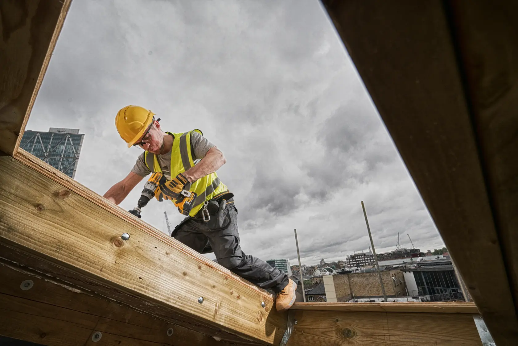 Ein Bauarbeiter mit gelbem Sicherheitshelm und Warnweste arbeitet mit einem DEWALT Akku-Bohrer an einem Holzbalken auf einer Baustelle im Freien, im Hintergrund ist die Stadt unter einem bewölkten Himmel zu sehen.
