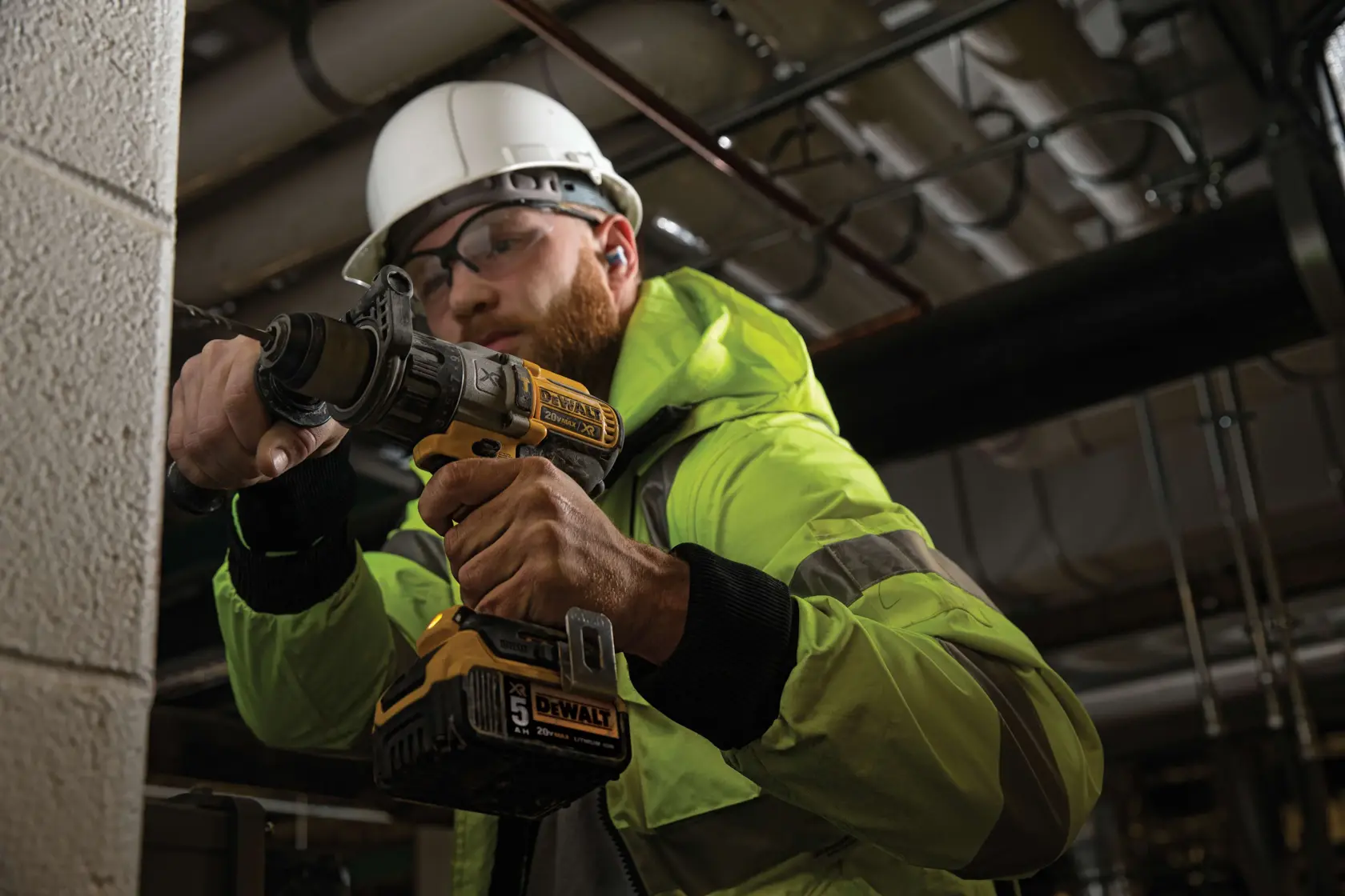 Una persona con chaqueta de alta visibilidad y casco de seguridad está utilizando un taladro atornillador inalámbrico DEWALT (modelo DCD996P2) para perforar una pared de concreto o ladrillo en un entorno industrial o de construcción. El producto y el agarre de la herramienta son claramente visibles.