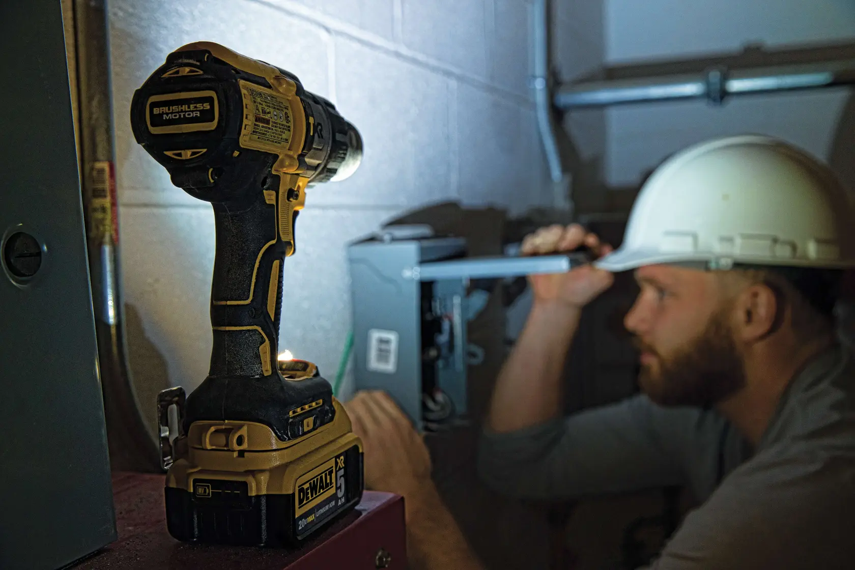 Un taladro inalámbrico DEWALT con motor sin escobillas está sobre una superficie en primer plano, mientras una persona con casco de seguridad trabaja en una caja eléctrica en el fondo. El rostro de la persona está difuminado para preservar la privacidad.