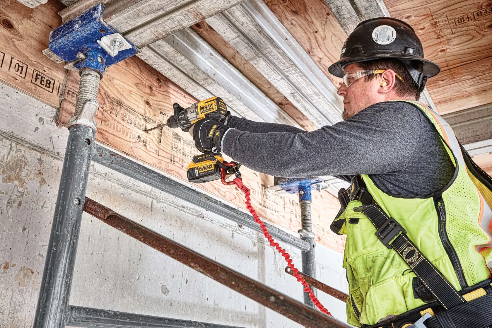 Un trabajador de la construcción con chaleco de seguridad y casco utiliza un taladro inalámbrico DEWALT (SKU: DCD996P2) para perforar vigas de madera en un sitio de construcción, de pie sobre un andamio.