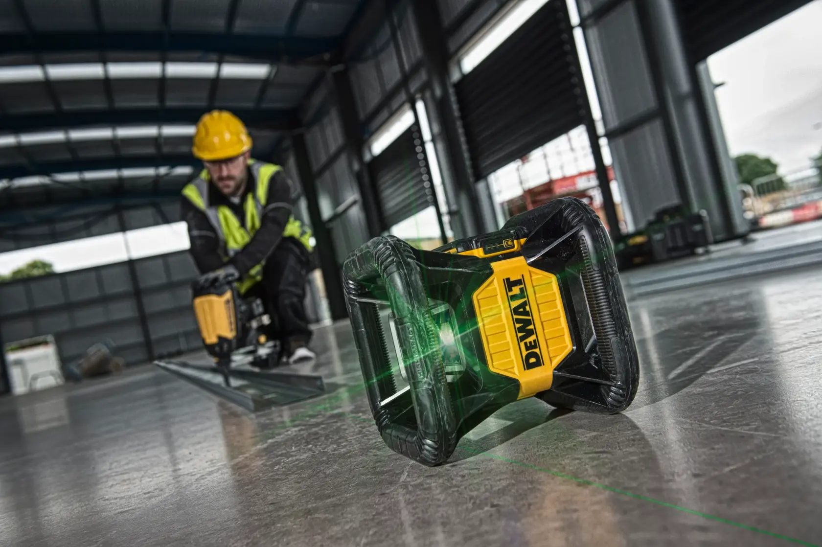 Nivel láser DEWALT proyecta línea verde en suelo de obra, trabajador de fondo con herramienta eléctrica.