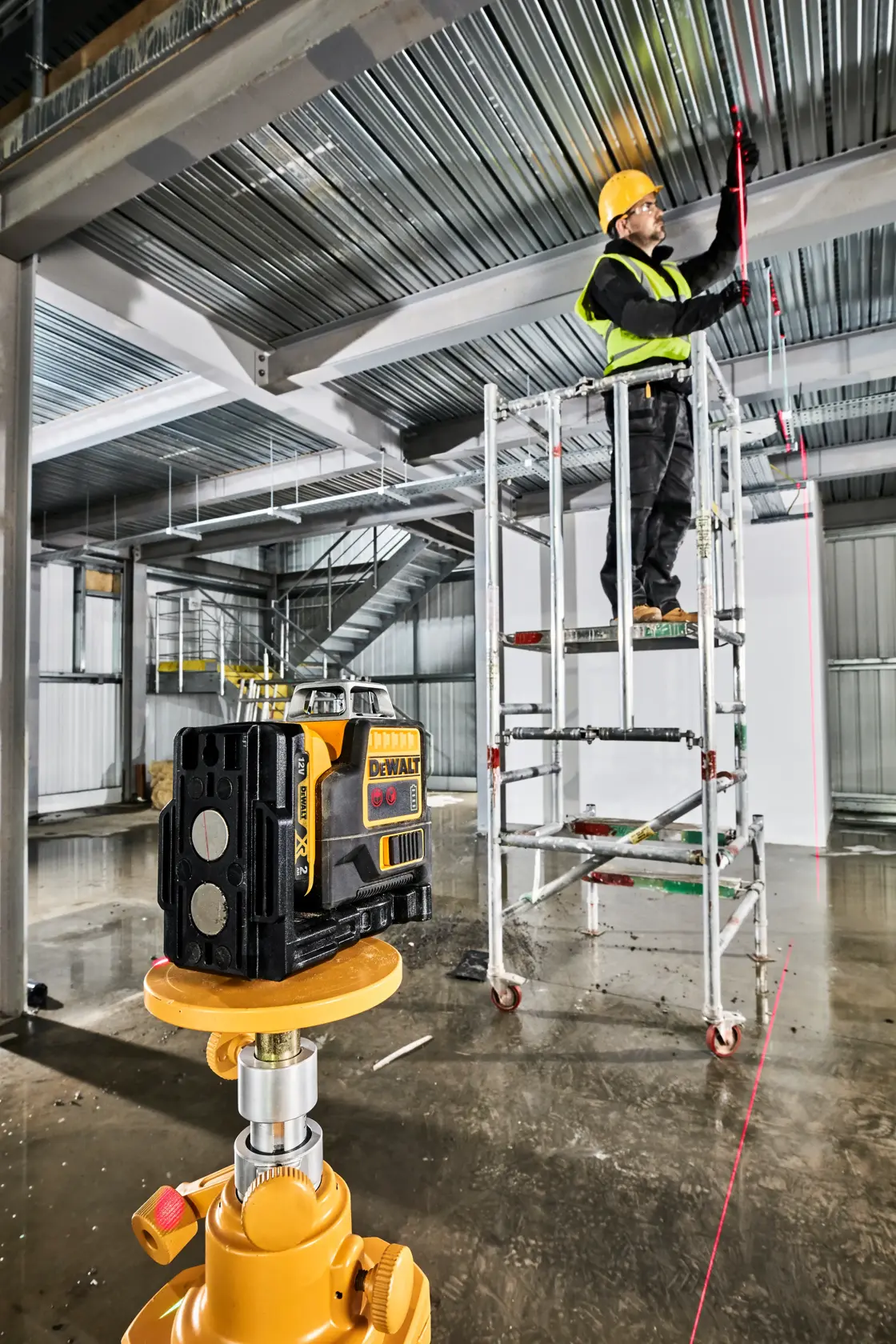 A DEWALT laser level device is set up on a tripod in the foreground of an industrial construction site. In the background, a worker wearing a yellow hard hat and high-visibility vest stands on scaffolding, marking a metal ceiling along the visible red laser line projected by the device. The area features metal beams and stairs, with construction materials visible throughout.