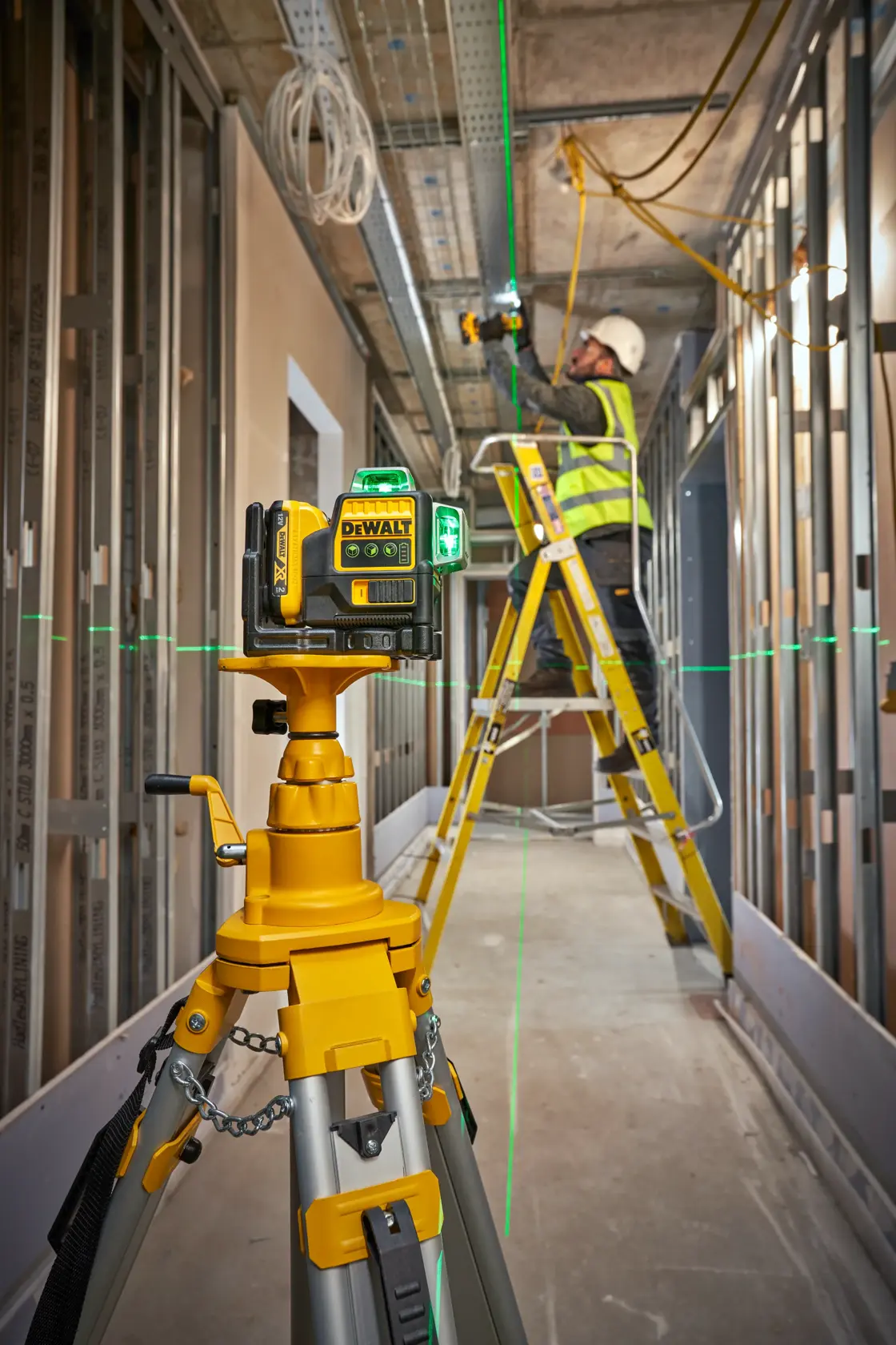 DEWALT Laser-Niveau auf Stativ projiziert grüne Linien, Arbeiter auf Leiter in Baustelle.