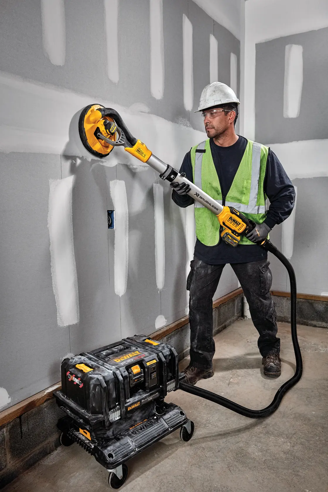 Un trabajador de la construcción con casco y chaleco de seguridad está lijando una pared de yeso con una lijadora inalámbrica para yeso de DEWALT, modelo DCE800T2. La lijadora está conectada mediante una manguera a un extractor de polvo portátil en el suelo.
