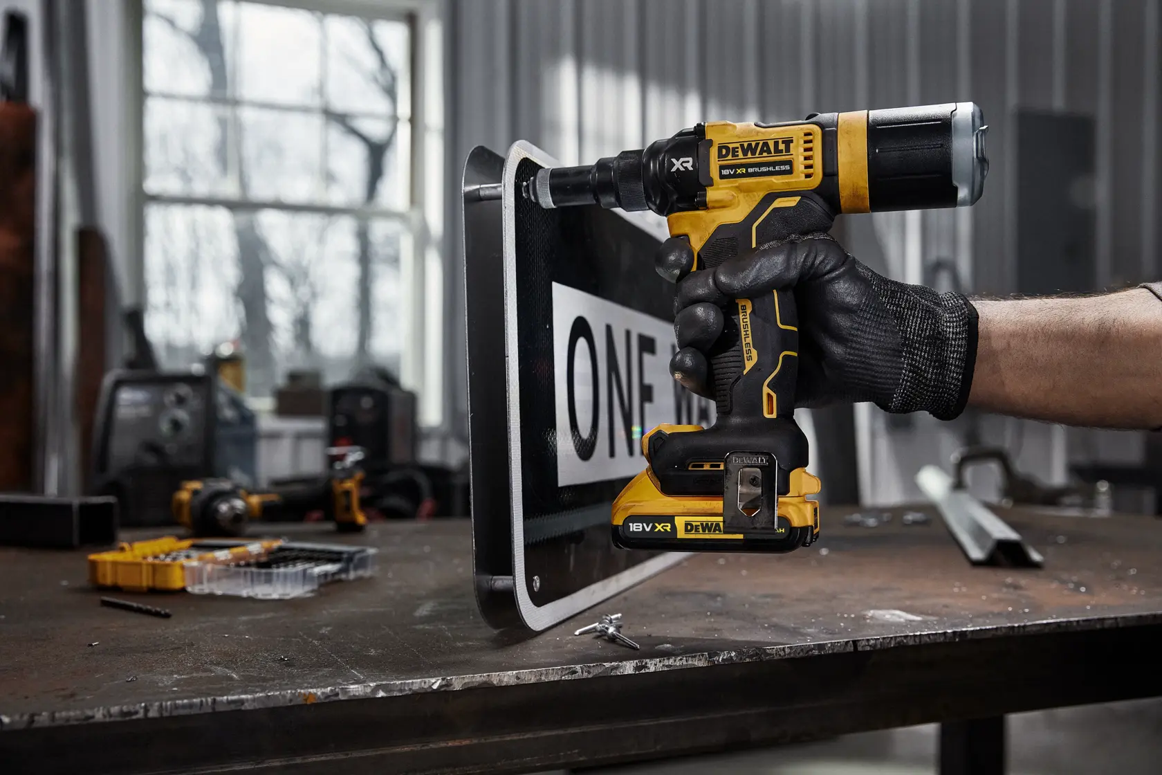 Una persona con un guante de trabajo negro utiliza un taladro inalámbrico DEWALT DCF403NT 18V XR para perforar una señal metálica sobre un banco de trabajo en un taller.