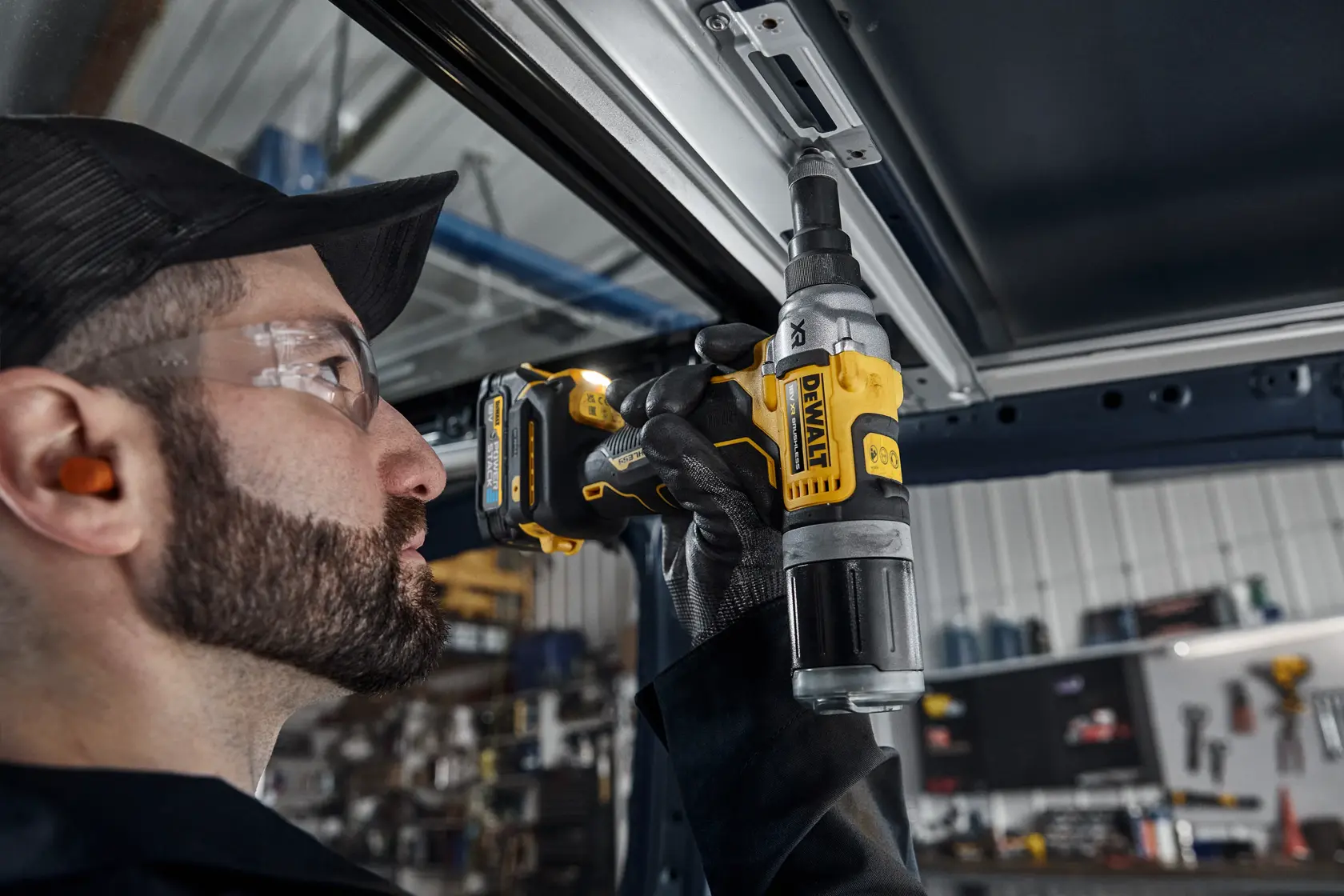 Una persona con protección auditiva y guantes utiliza una herramienta eléctrica inalámbrica DEWALT DCF414N para fijar o aflojar un componente metálico en un entorno interior de taller o industrial.