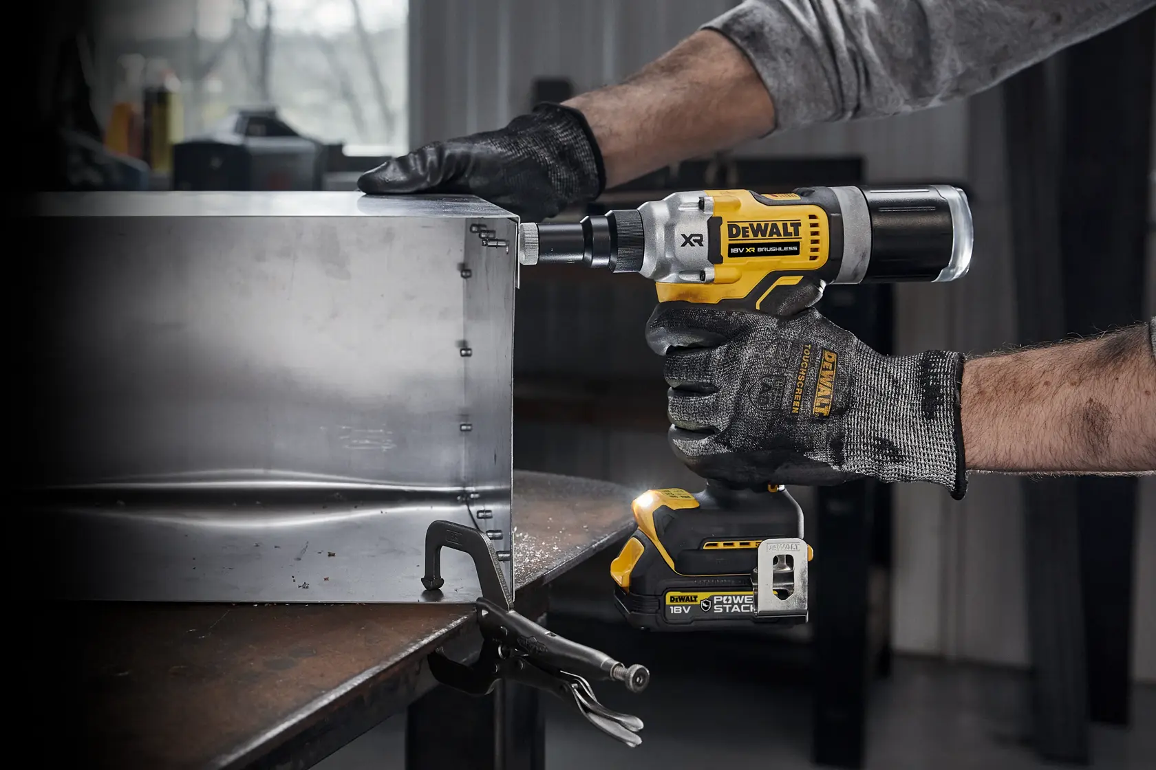 Una persona con guantes de trabajo utiliza una remachadora inalámbrica DEWALT DCF414NT de 18V para fijar una lámina de metal en un taller industrial.