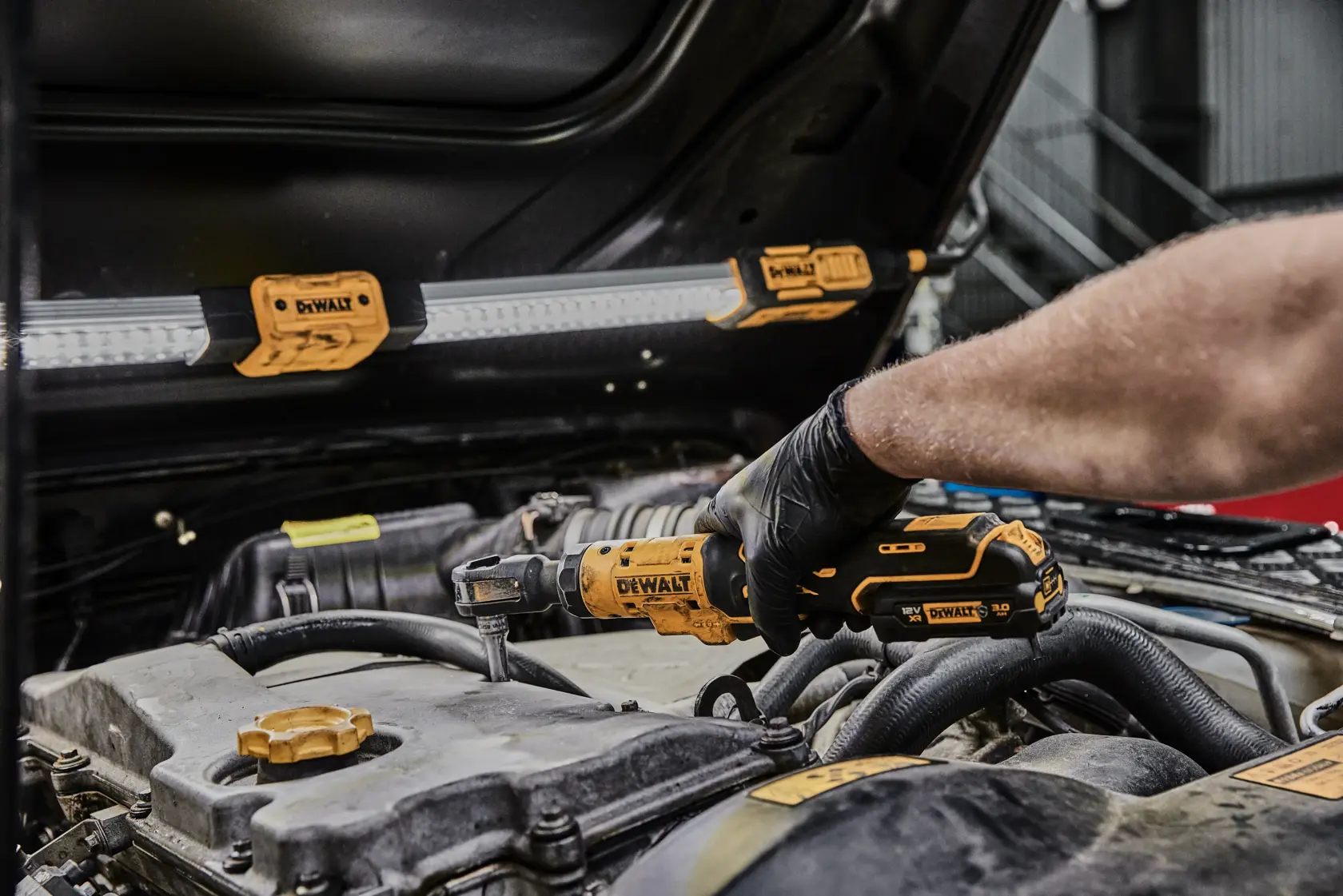 Eine Person mit einem schwarzen Handschuh verwendet ein gelbes DEWALT Akku-Ratschenwerkzeug (DCF503L1G), um an einem Motor unter der Motorhaube eines Autos zu arbeiten. Mehrere DEWALT LED-Arbeitsleuchten sind zur Beleuchtung an der Unterseite der Motorhaube angebracht.