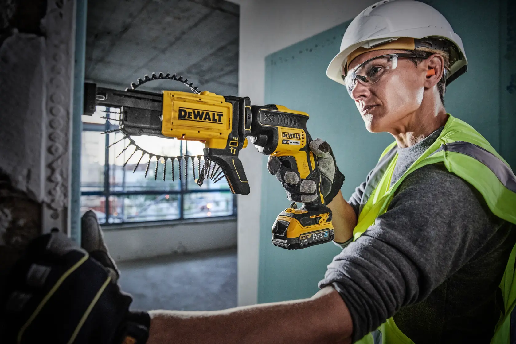 A construction worker wearing a safety helmet and vest uses a DEWALT DCF620E1K cordless drywall screw gun to fasten drywall sheets. The tool features an attached screw magazine for quick and efficient installation.