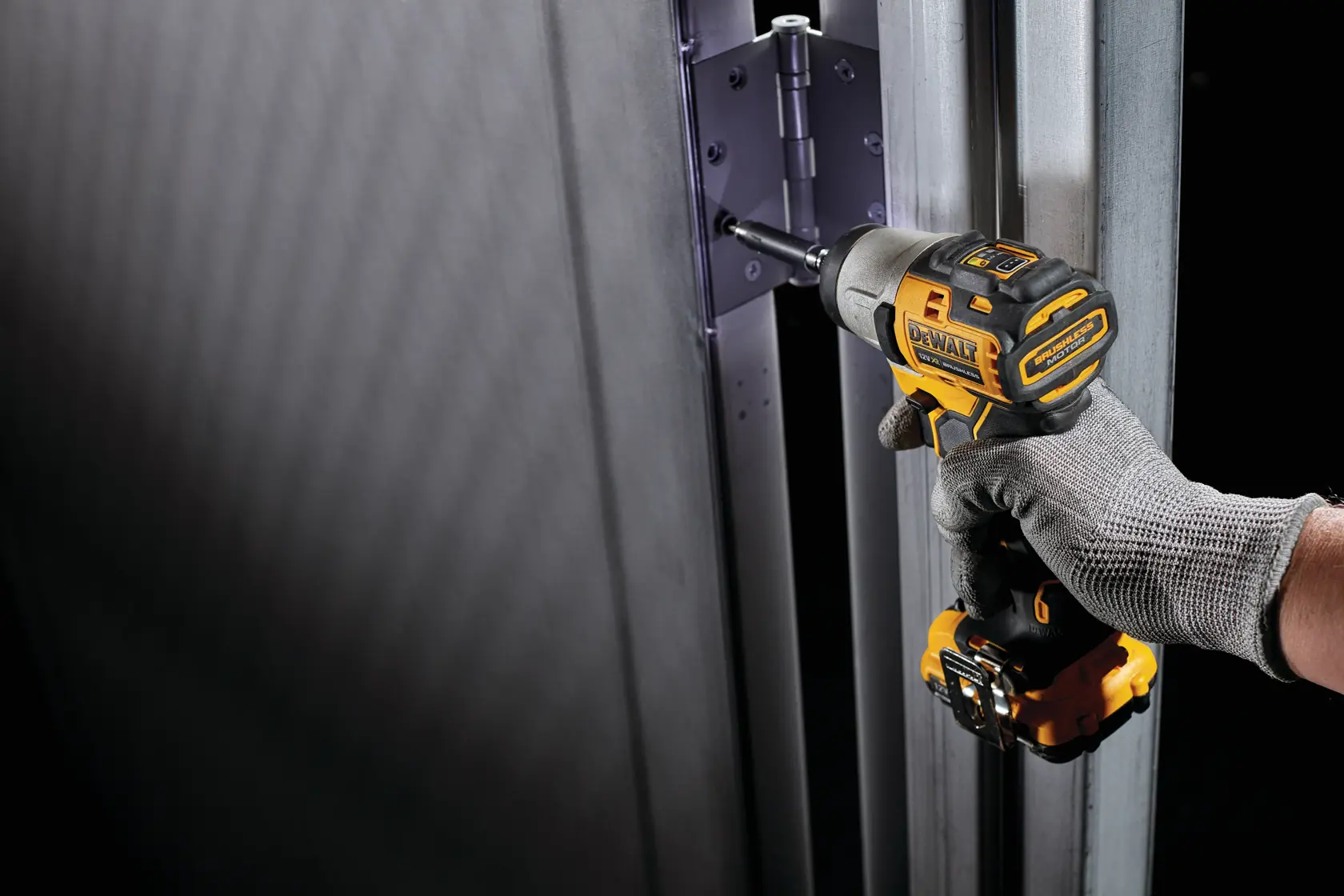 Una persona con un guante de trabajo gris utiliza un atornillador de impacto inalámbrico DEWALT para instalar o apretar tornillos en una bisagra de puerta metálica.