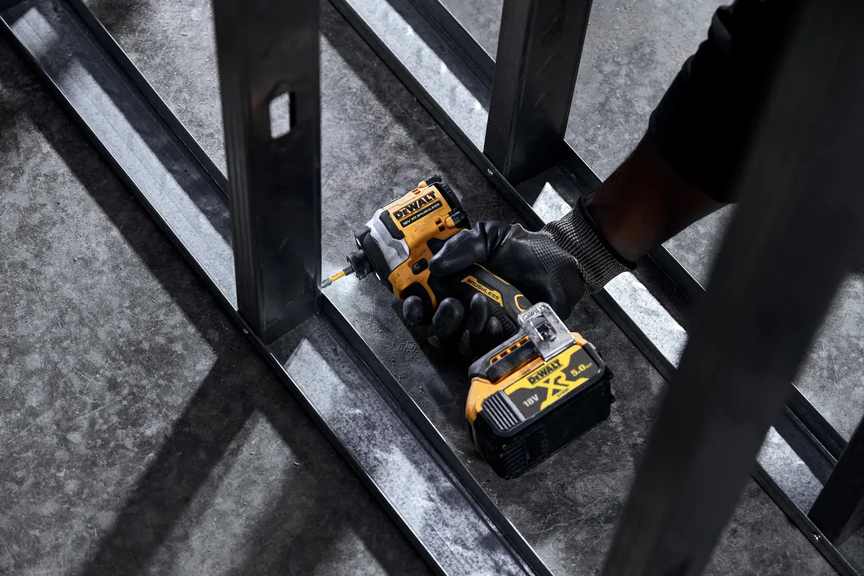 Mano enguantada usa un atornillador de impacto DEWALT inalámbrico sobre estructura metálica.