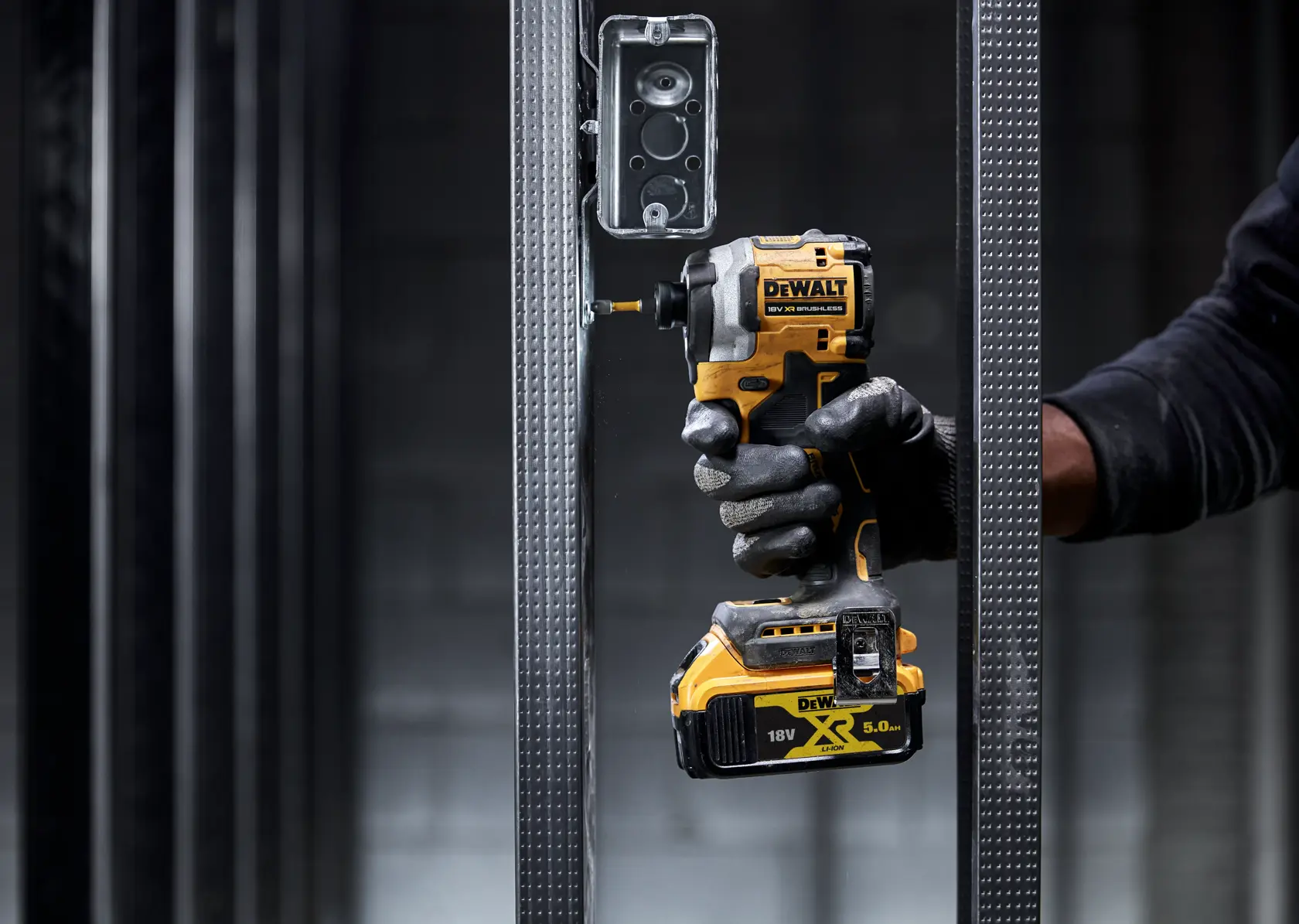 Mano con guante usa DEWALT DCF850NT para atornillar en marco metálico junto a una caja eléctrica.