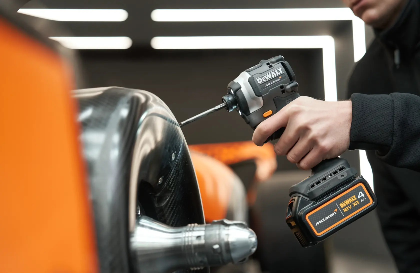 A person uses a DEWALT cordless impact driver on a metallic and carbon fiber surface beside orange details.