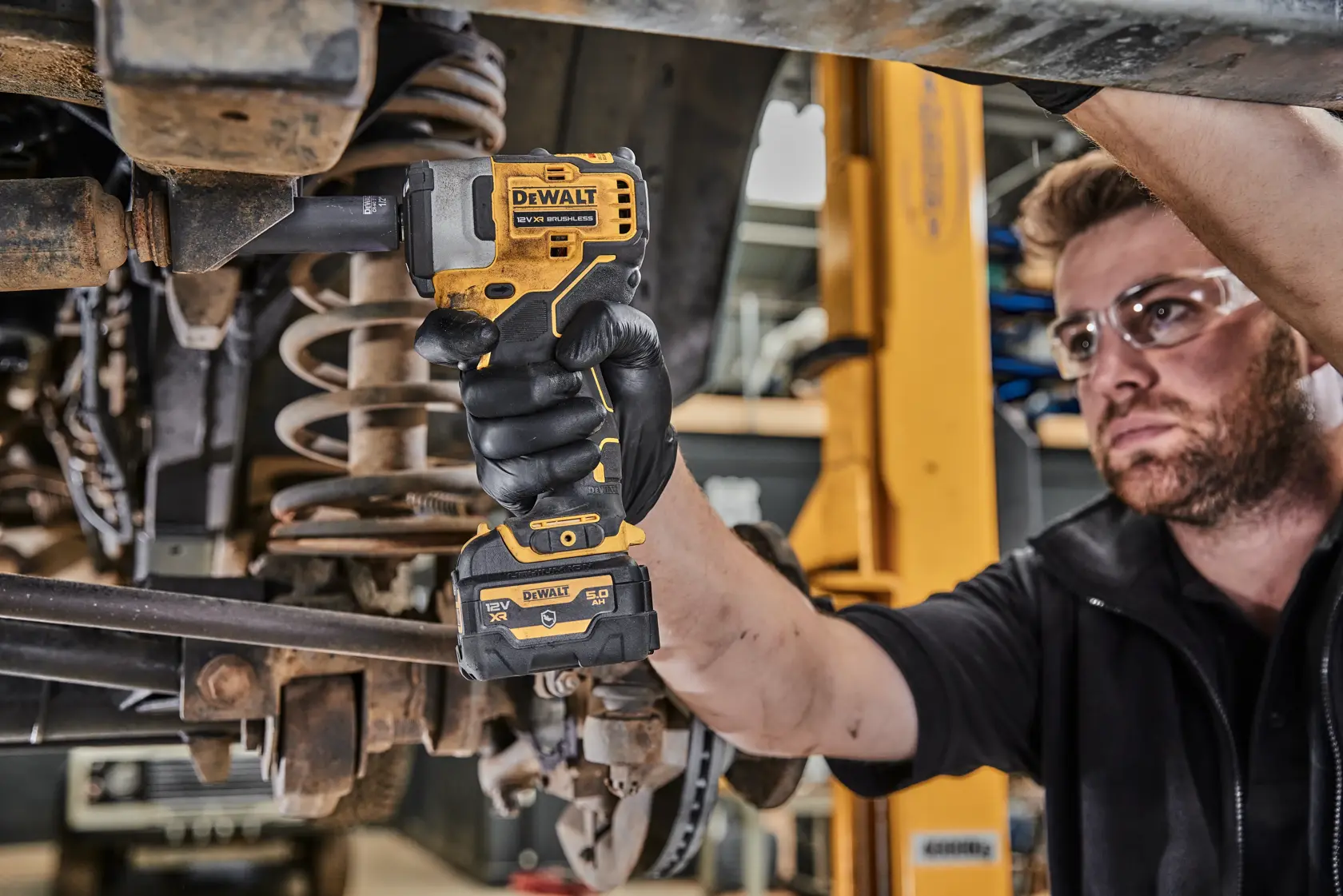 Eine Person verwendet einen kabellosen Schlagschrauber von DEWALT (DCF901P1G), um an der Unterseite eines Fahrzeugs in einer Werkstatt zu arbeiten. Die Person trägt schwarze Handschuhe und das Werkzeug wird an einer Schraube angesetzt.