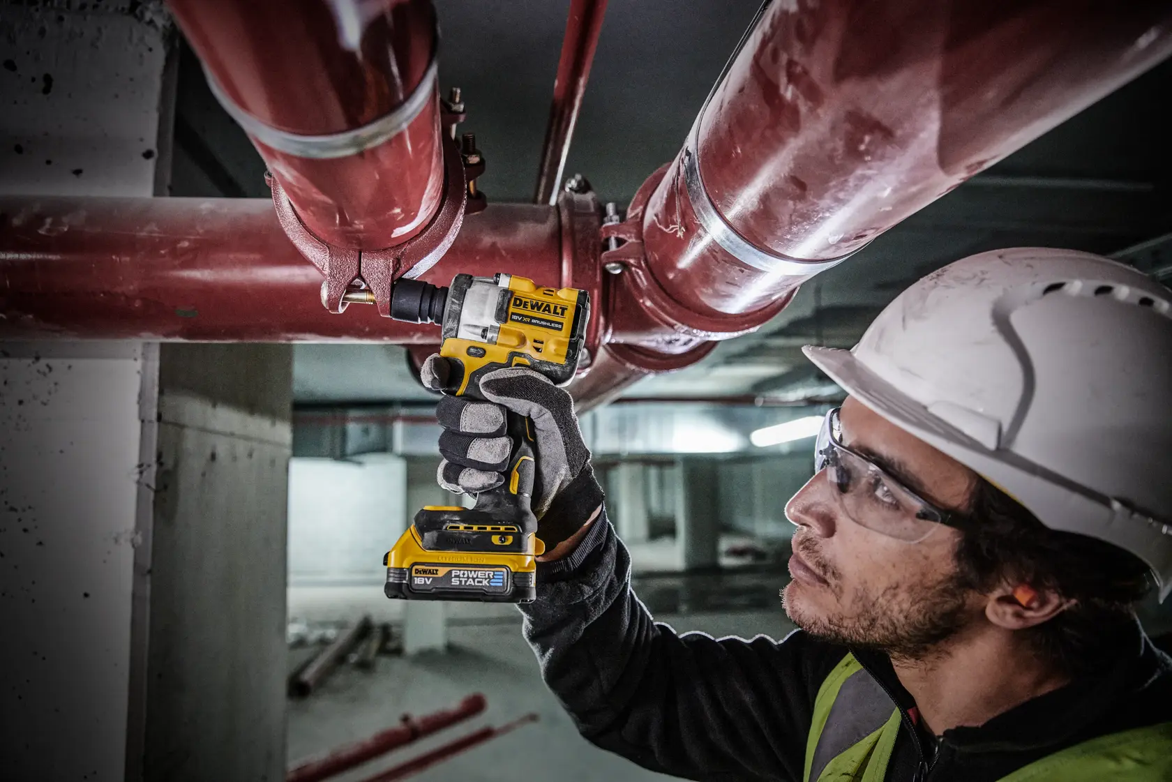 A person wearing a safety helmet and gloves uses a DEWALT cordless impact wrench (model DCF921E2T) to tighten a bolt on a large red pipe in an industrial setting. The person's face is blurred for privacy.