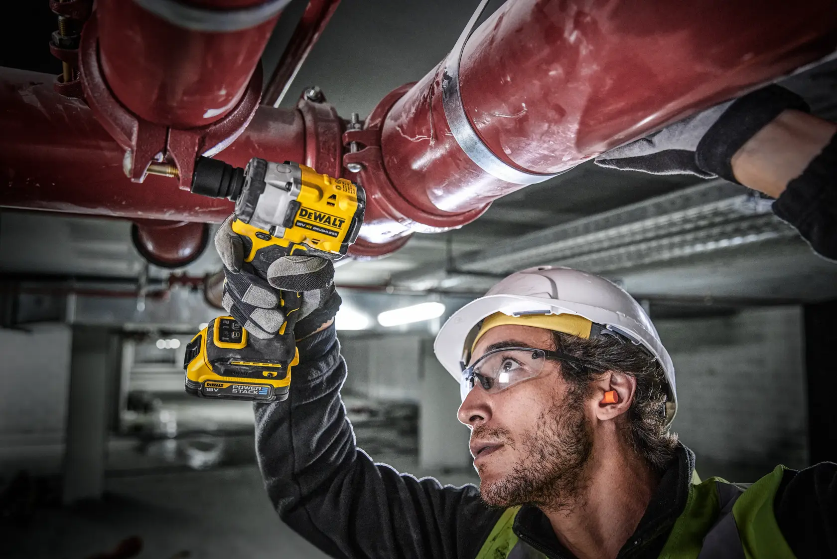 A person wearing safety gear uses a DEWALT cordless impact wrench (model DCF921E2T) to work on large red industrial pipes in an indoor setting. The person's face is blurred.
