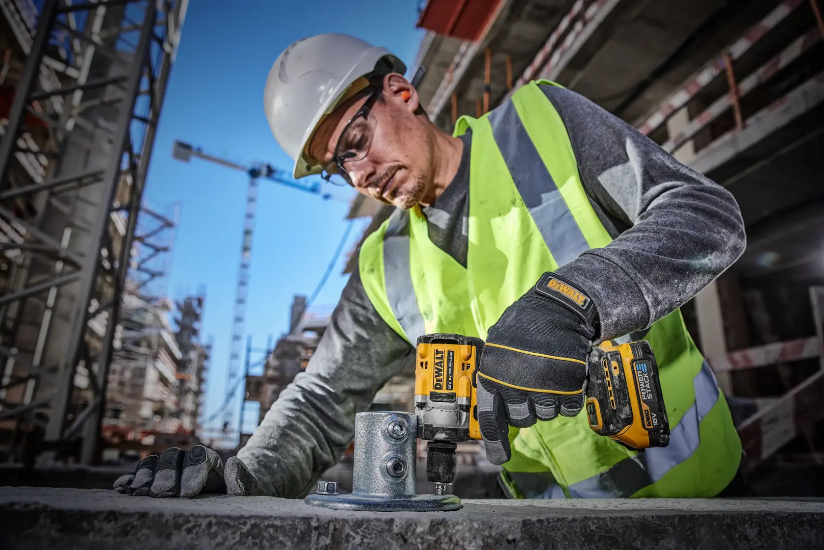 A construction worker wearing a safety helmet, high-visibility vest, and gloves is using a DEWALT DCF921E2T cordless impact wrench to secure bolts on a metal structure at a busy construction site.