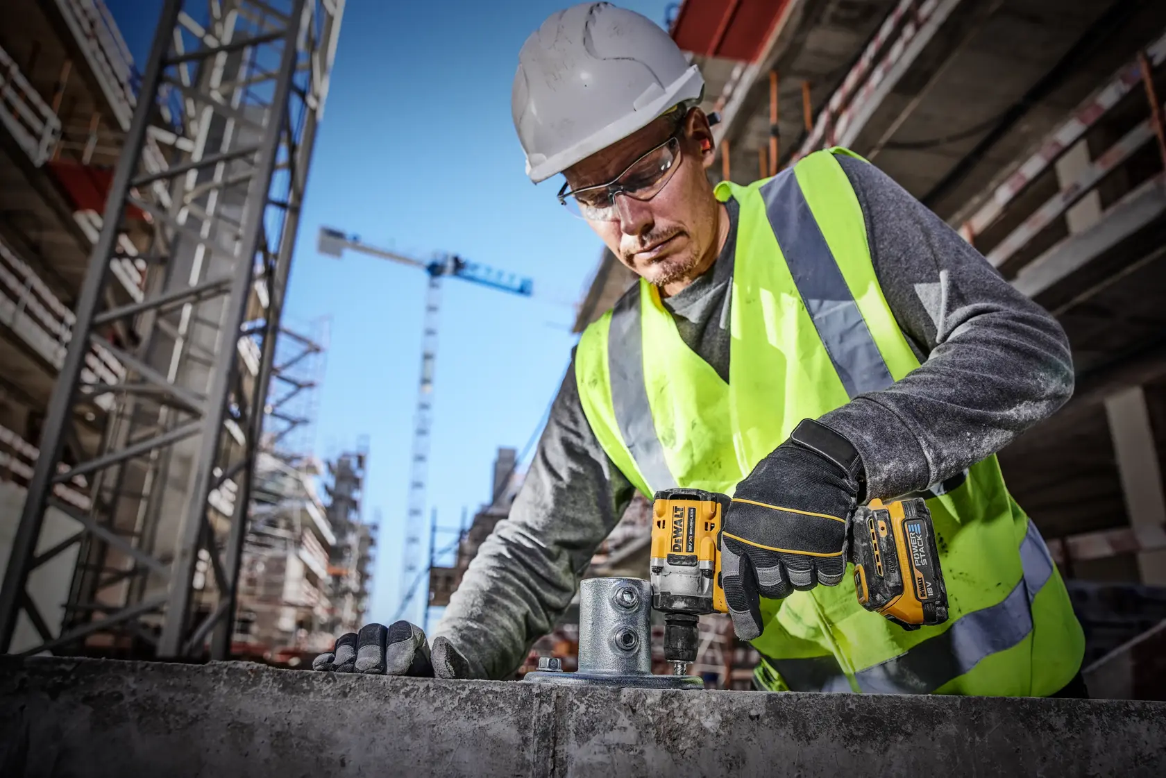 A construction worker wearing a high-visibility vest and safety helmet uses a DEWALT DCF921E2T power tool on a building site. Several cranes and unfinished buildings are visible in the background.