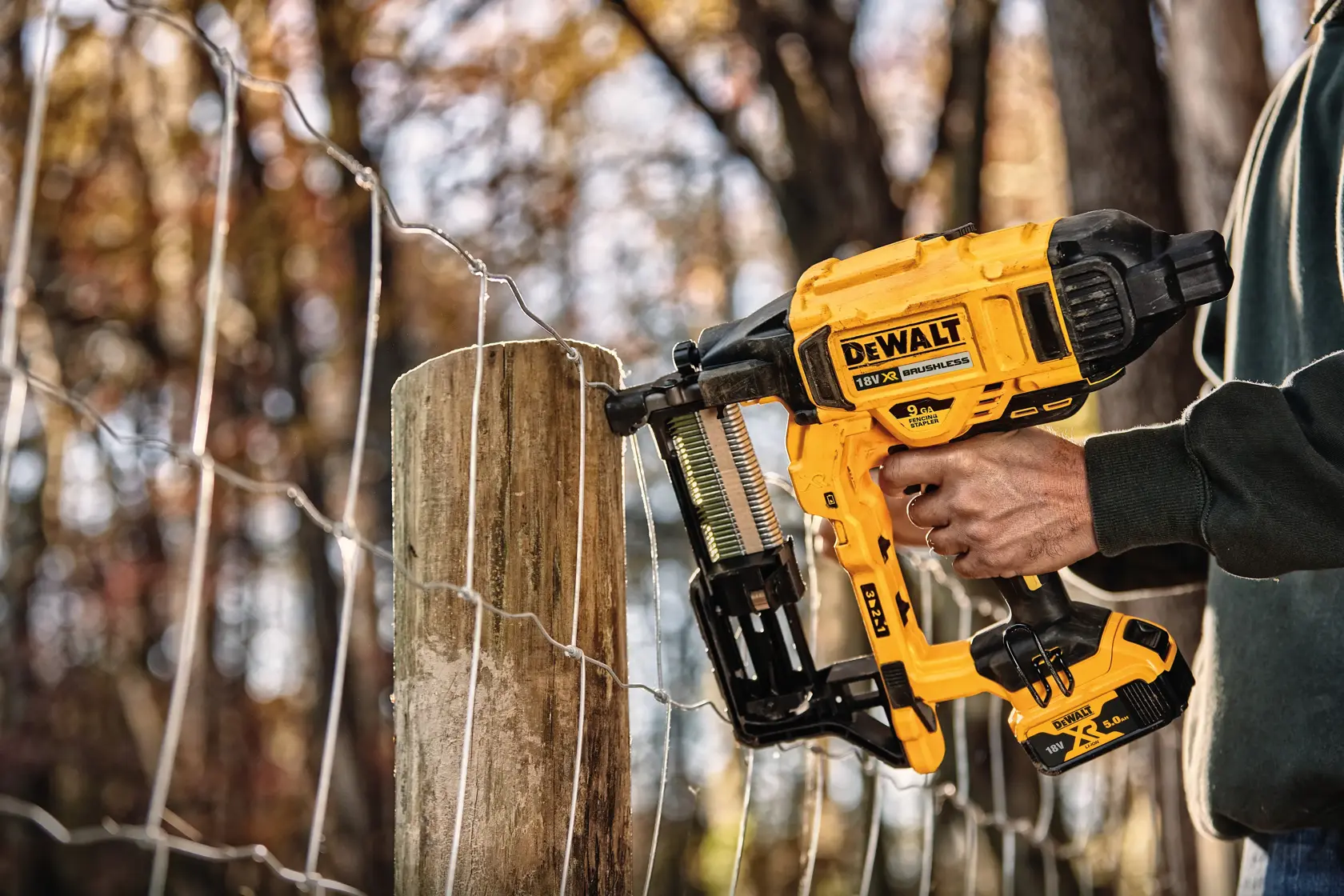 Una persona utiliza una grapadora para cercas DEWALT 18V inalámbrica para fijar una malla de alambre a un poste de madera al aire libre.