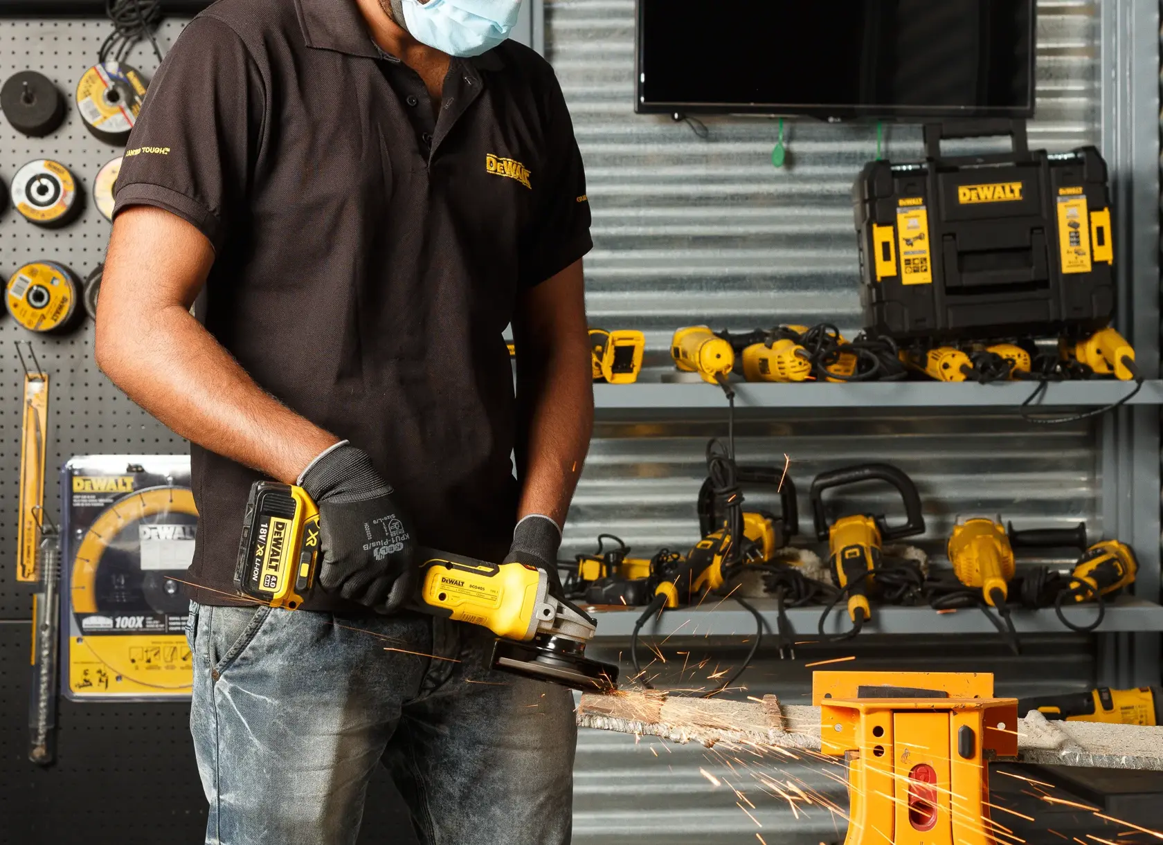 Persona usa amoladora DEWALT sin cable en metal, con guantes y mascarilla. Herramientas en el fondo visibles.