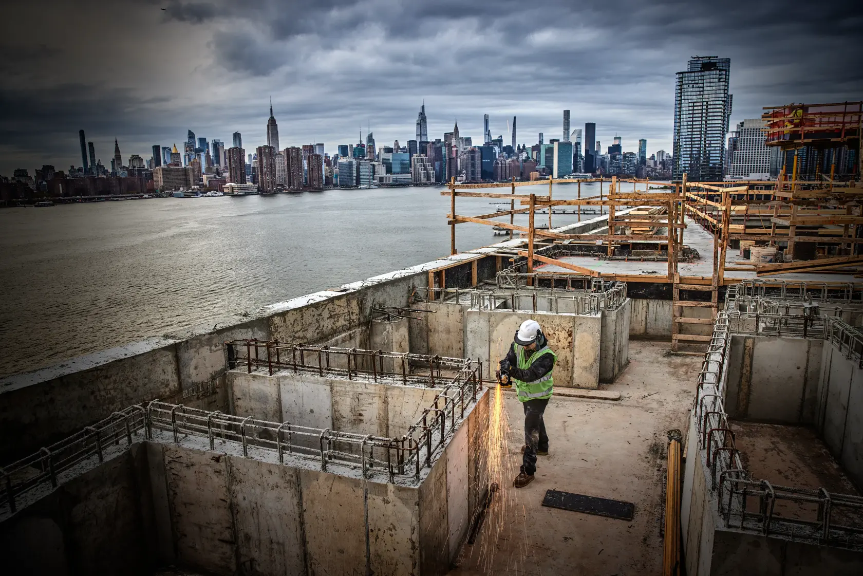 Trabajador usa herramienta en concreto en obra cerca del agua, fondo de skyline urbano.
