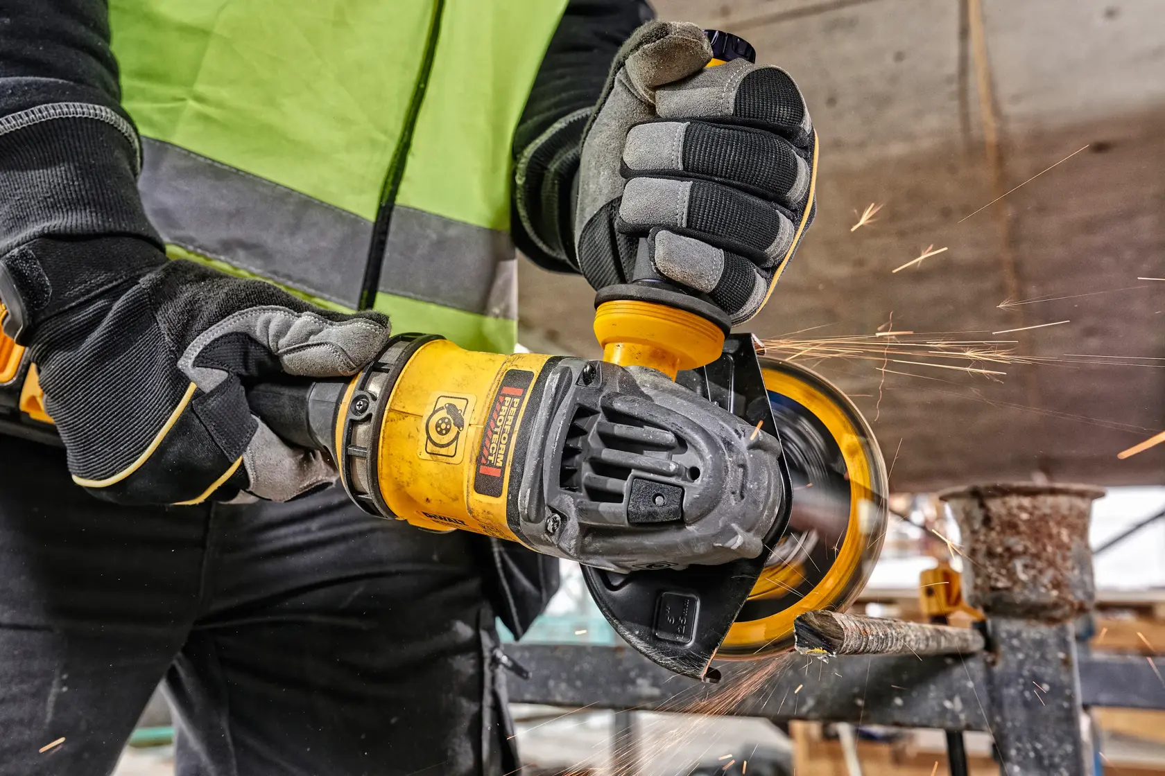 Worker uses DEWALT angle grinder to cut metal, sparks flying, wearing gloves and safety vest at construction site.