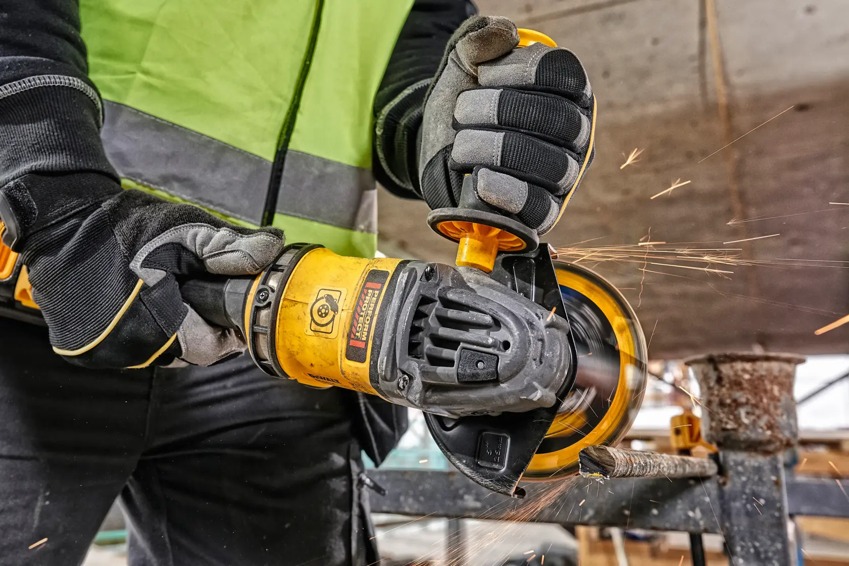 Trabajador usa amoladora DEWALT amarilla para cortar metal, con guantes y chaleco. Hay chispas visibles.