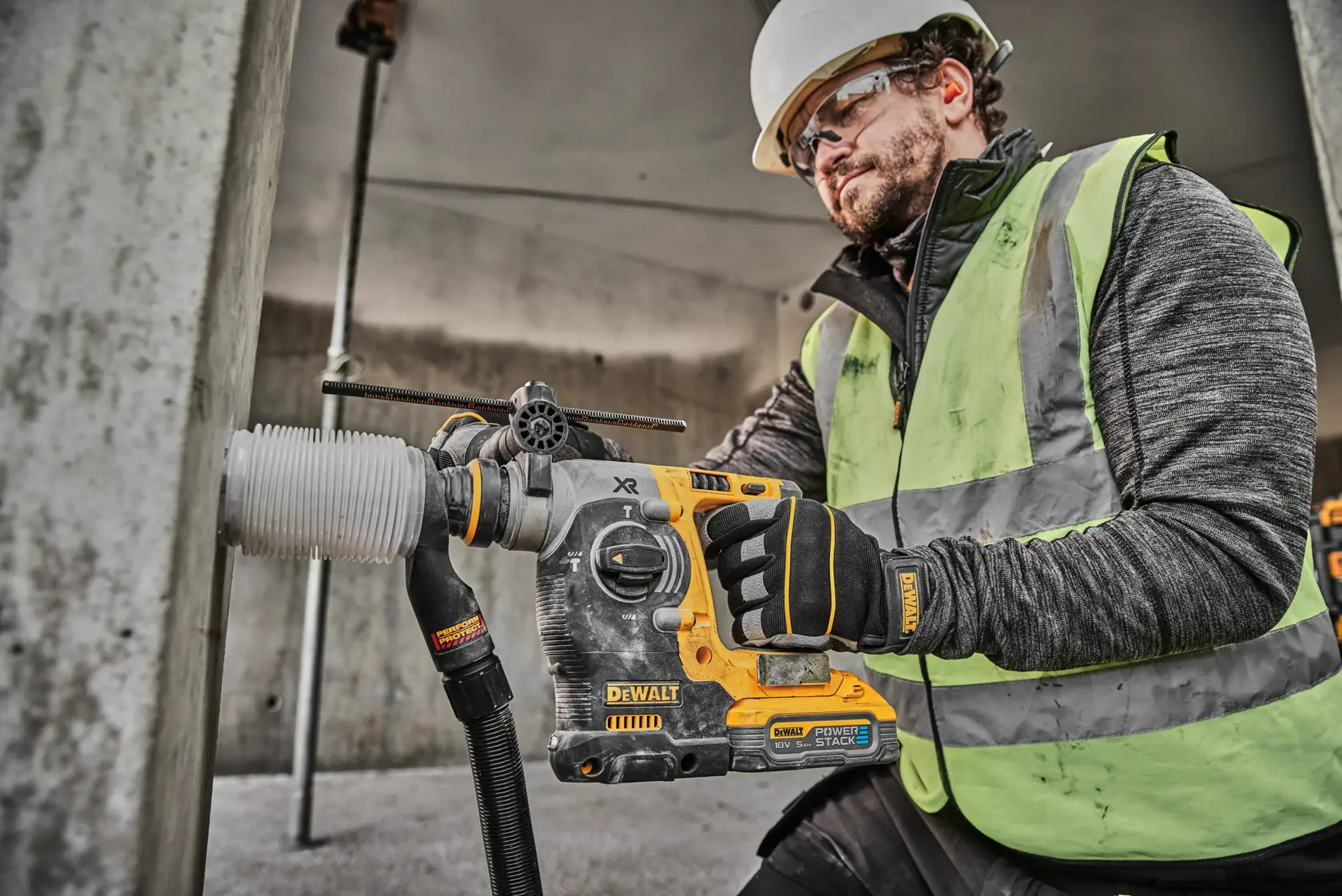 Un trabajador de la construcción con casco de seguridad y chaleco reflectante utiliza un martillo rotativo inalámbrico DEWALT DCH273H2T para perforar una columna de hormigón. El trabajador también lleva guantes DEWALT.