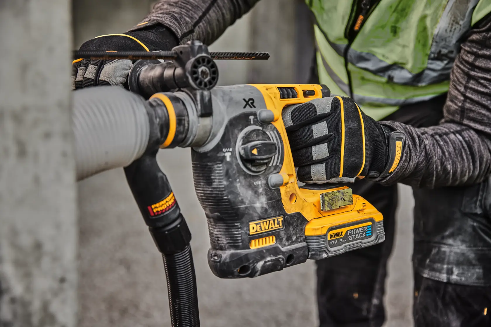 Primer plano de un trabajador usando un taladro rotatorio inalámbrico DEWALT con batería POWERSTACK para perforar concreto, usando guantes y ropa de protección.