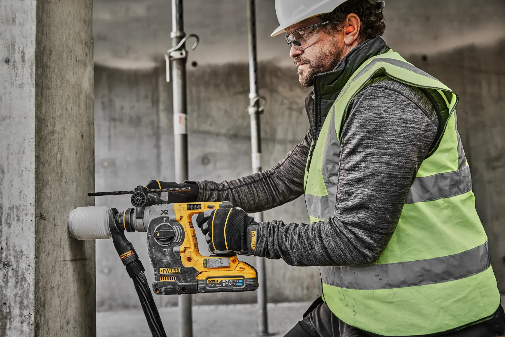 Una persona con chaleco de seguridad y guantes está utilizando un martillo rotativo inalámbrico DEWALT DCH273H2T para perforar un pilar de concreto en un sitio de construcción. La cara del usuario está desenfocada por motivos de privacidad.