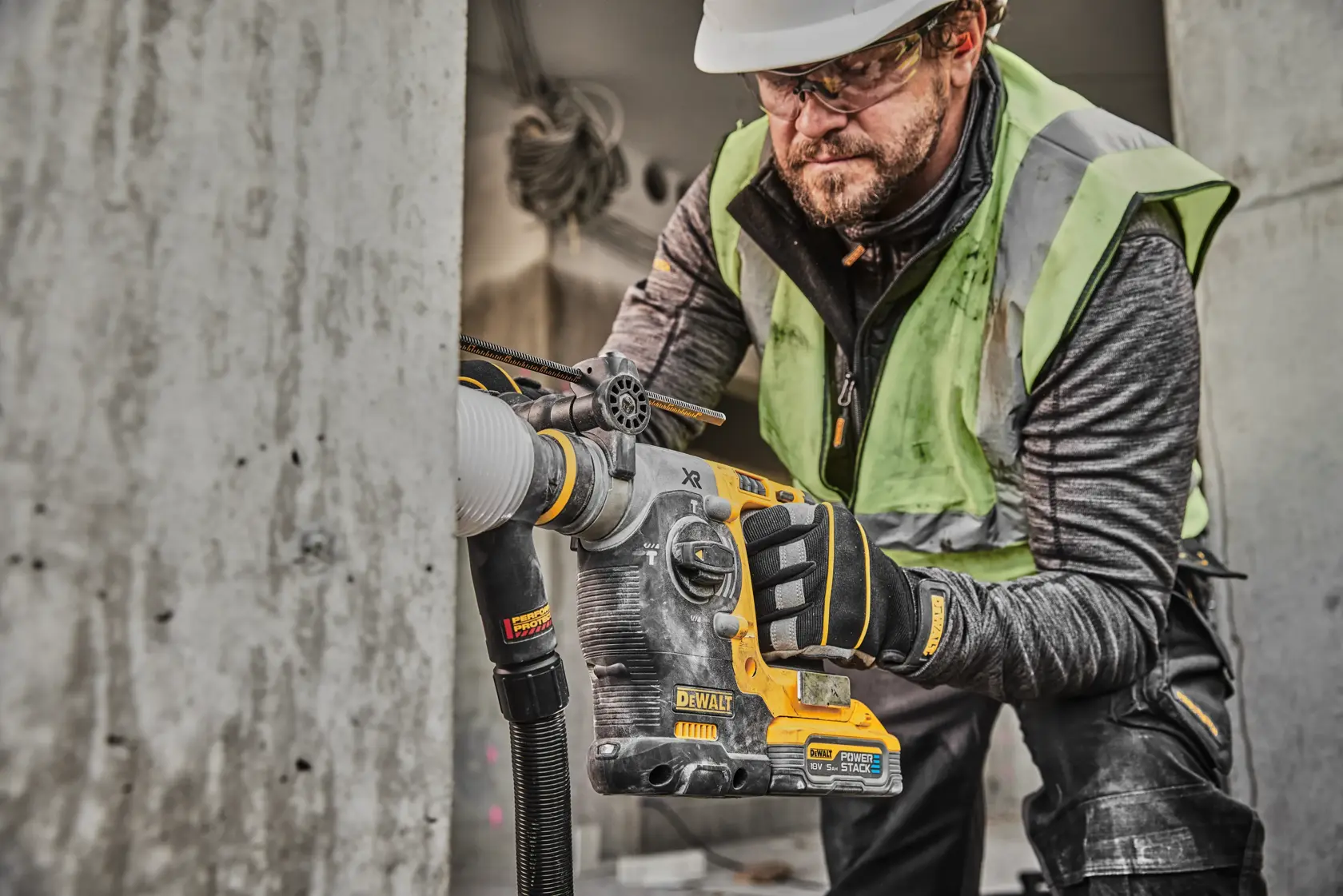 Un trabajador de la construcción usa un martillo rotativo DEWALT DCH273H2T con extracción de polvo para perforar concreto. El trabajador lleva guantes de seguridad, chaleco reflectante y casco. El taladro es DEWALT y presenta una batería PowerStack.