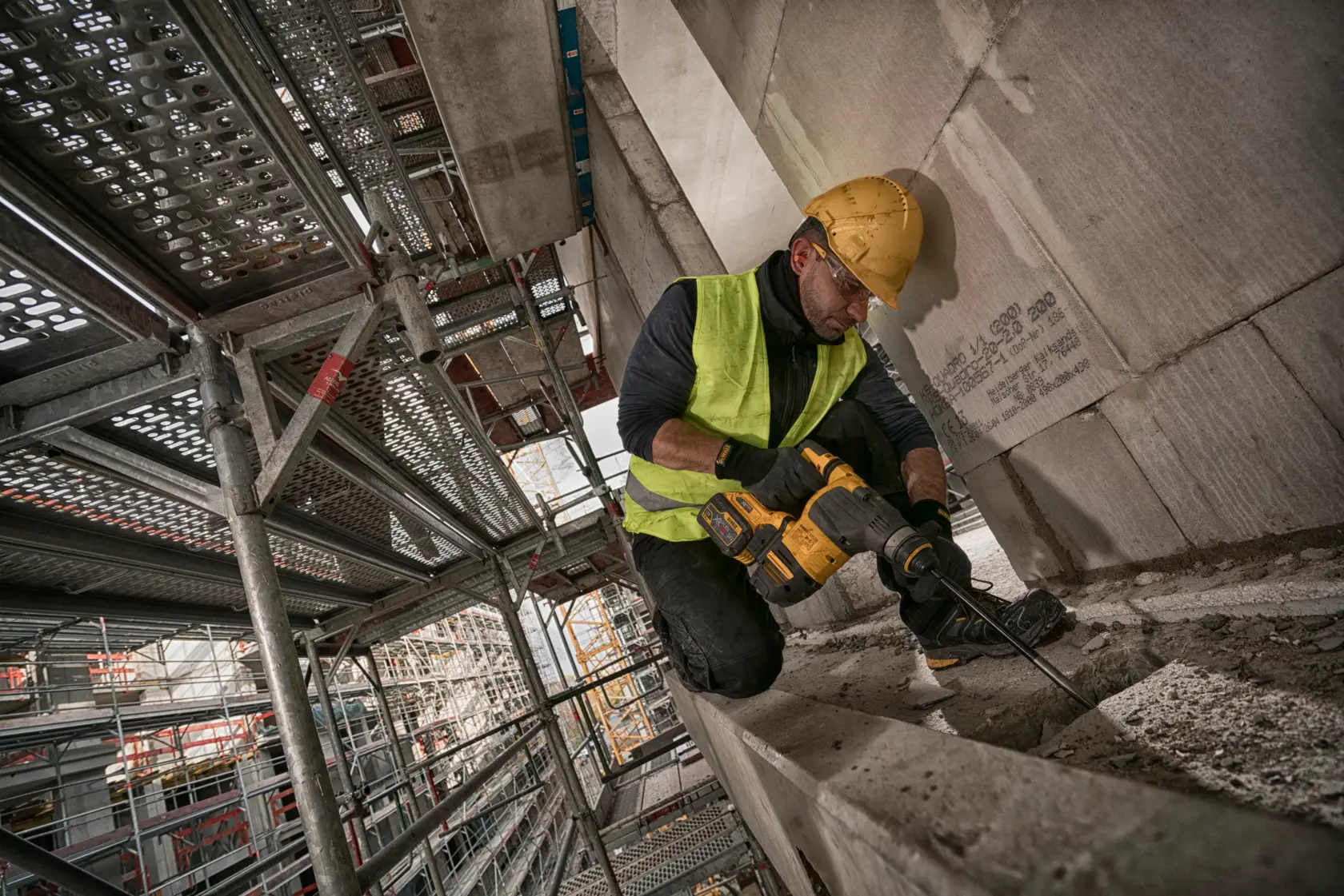 Trabajador usa un martillo rotativo DEWALT en obra, con andamios de fondo.