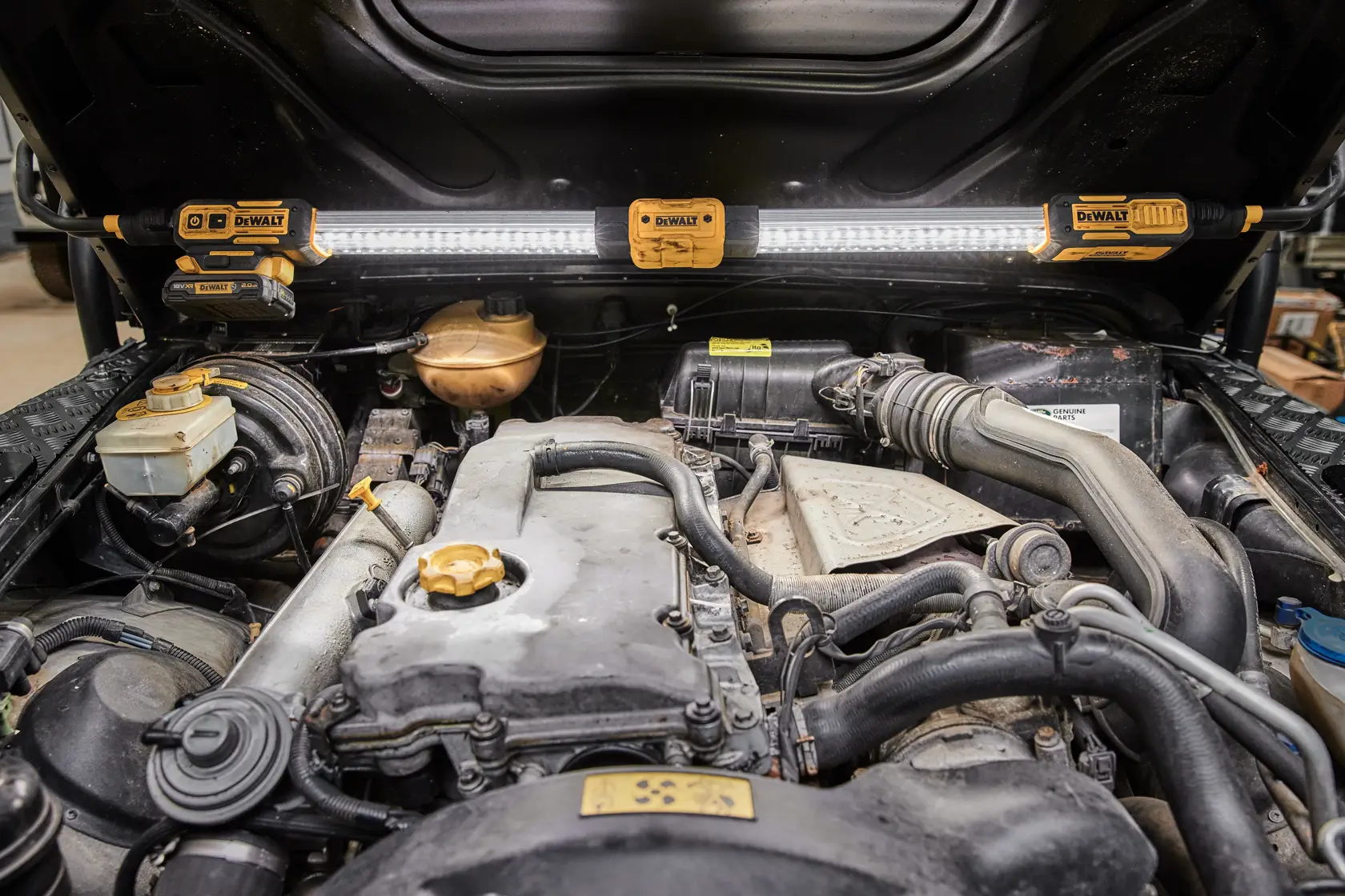 Una lámpara de trabajo LED DEWALT DCL045 está montada bajo el capó abierto de un vehículo, iluminando el compartimiento del motor para mejorar la visibilidad al trabajar.